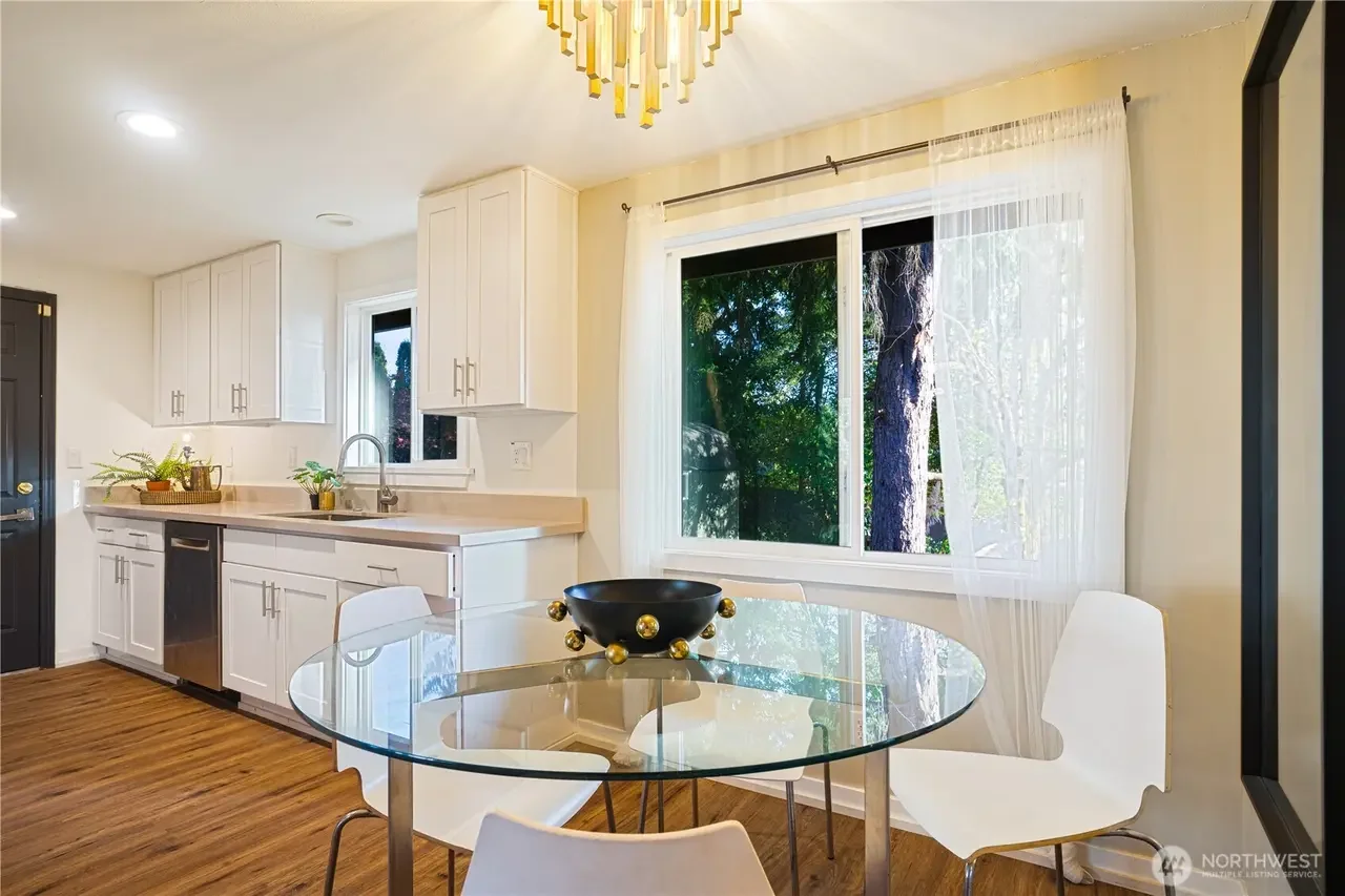 A bright kitchen with white cabinets, a glass dining table with white chairs, and large windows with sheer curtains showing trees outside.