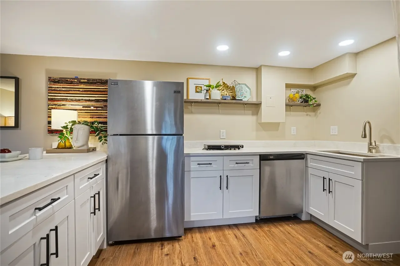 Kitchen with white cabinets, stainless steel refrigerator, dishwasher, beige walls, wooden floor, and open shelving with decor and plants.