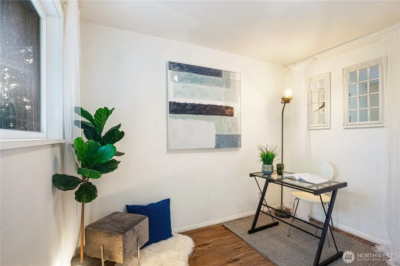 A small, minimalist home office with white walls, a window with sheer curtains, a black desk with a white chair, a tall floor lamp, a potted plant, a stool, a blue pillow, a white furry rug, and framed windows