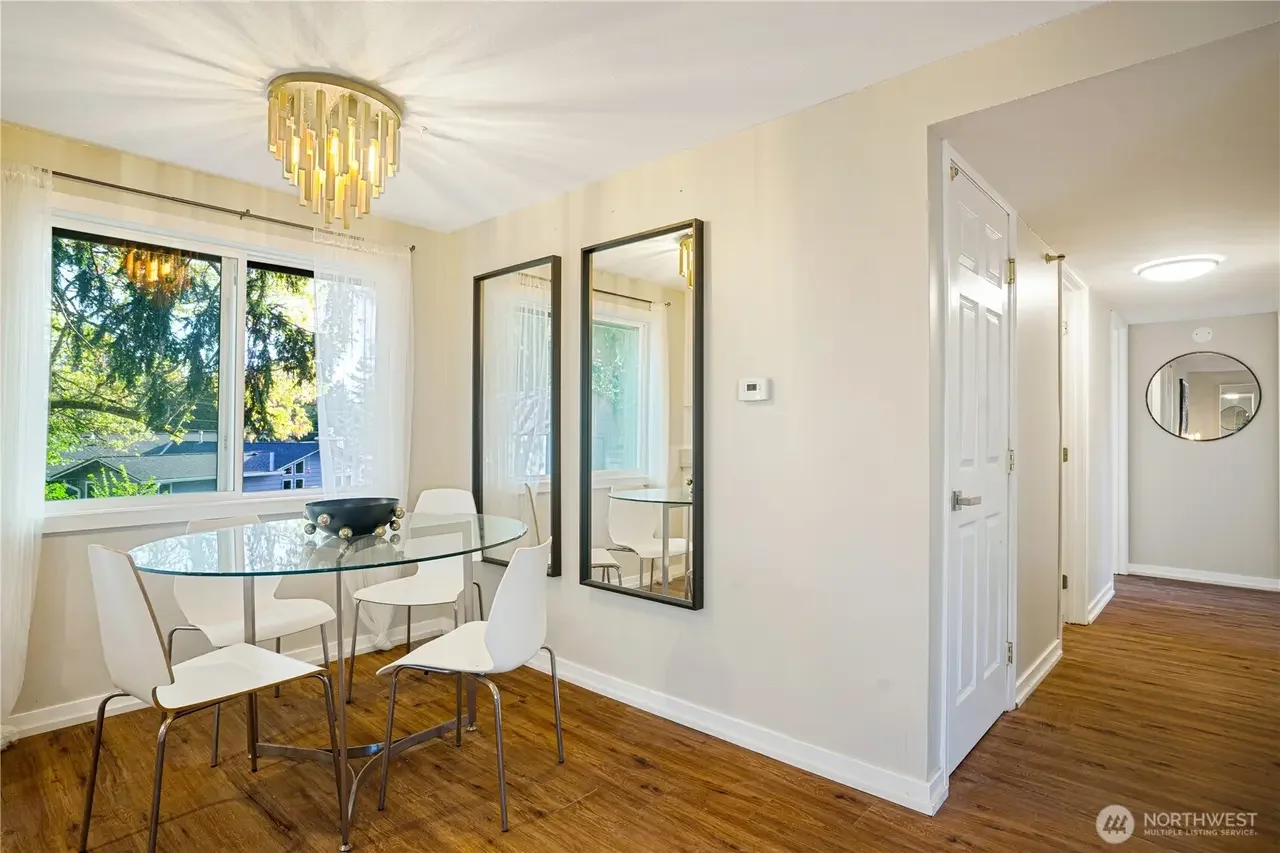 Dining area with a glass table, four white chairs, a large window with sheer curtains, and a modern gold chandelier. There are two tall mirrors on the wall, a thermostat, and a hallway with a round mirror on the far wall.