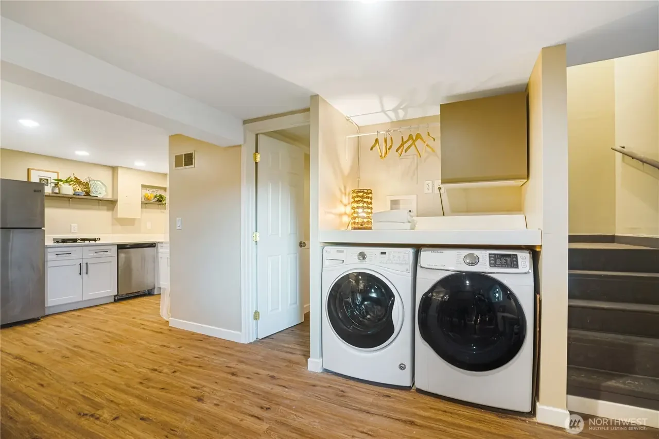 Laundry area with a washing machine and dryer beneath a shelf, next to a staircase in a kitchen space with wooden floors and white cabinets.