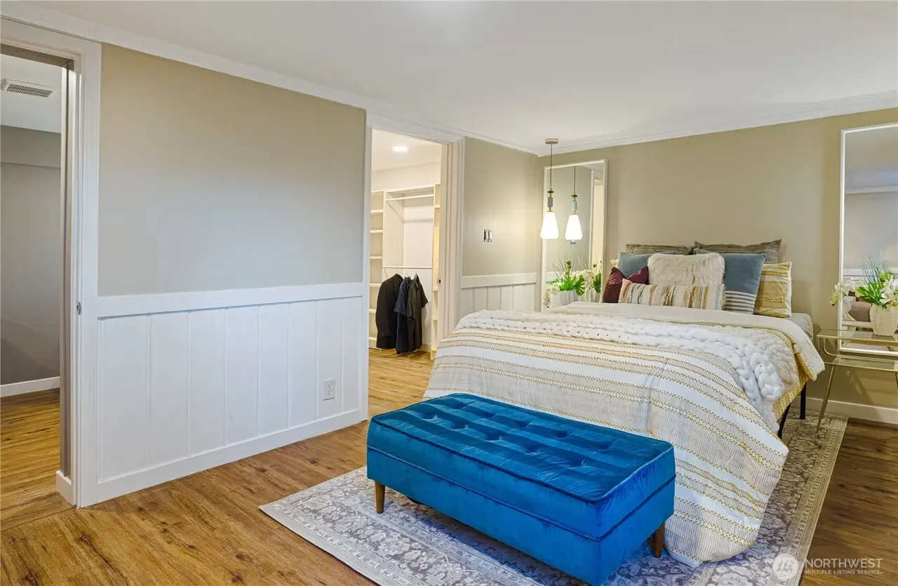 A bedroom with a bed, blue ottoman, and nightstand, featuring neutral walls, hardwood flooring, and closet in the background.