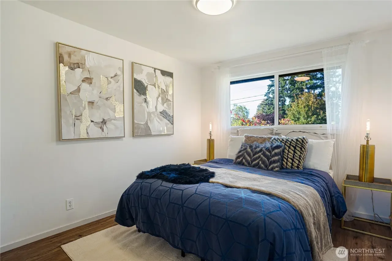 Bedroom with white walls, wooden floor, and a large window showing trees outside. The bed has white pillows, a blue quilt, and decorative pillows. There are two gold bedside tables with lamps, and abstract wall art hangs above the bed.