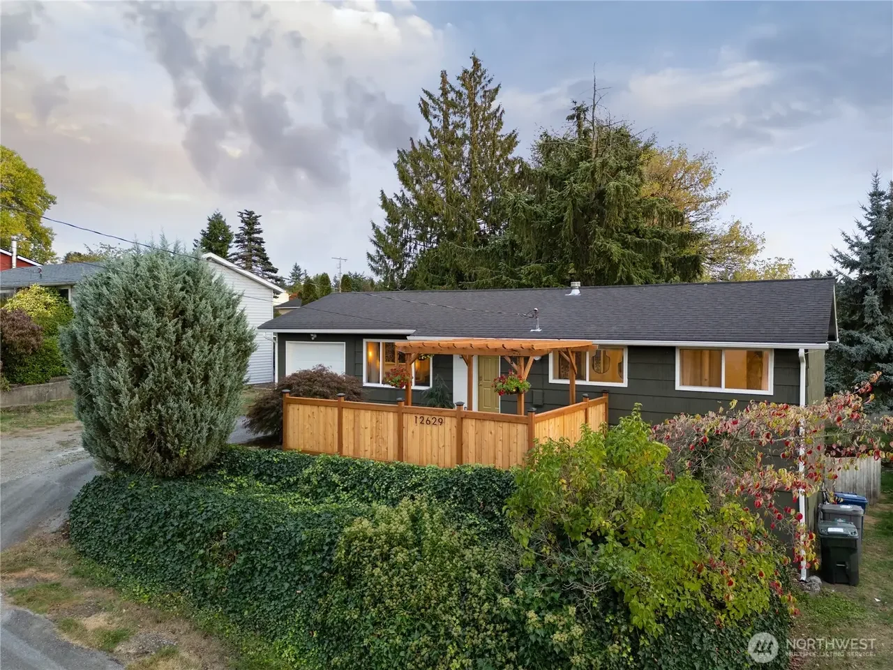 A house with a dark gray exterior, front porch with wooden railing and pergola, surrounded by green bushes and trees, under a partly cloudy sky.