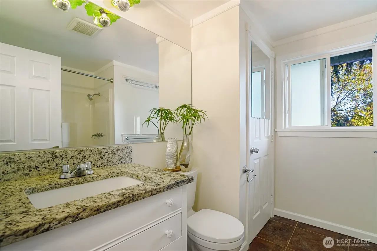 Bathroom with granite countertop, white sink, mirror, and windows showing trees outside.