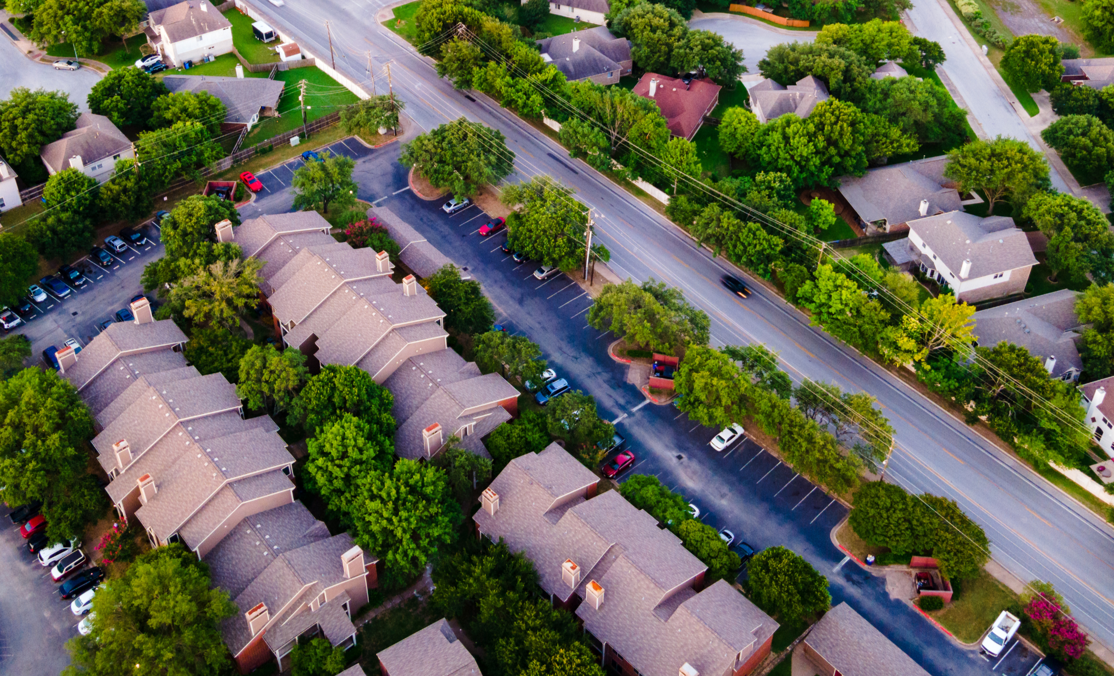 Aerial view of a residential neighborhood with multi-story townhouses, trees, parking lots, and a main road with moving vehicles.