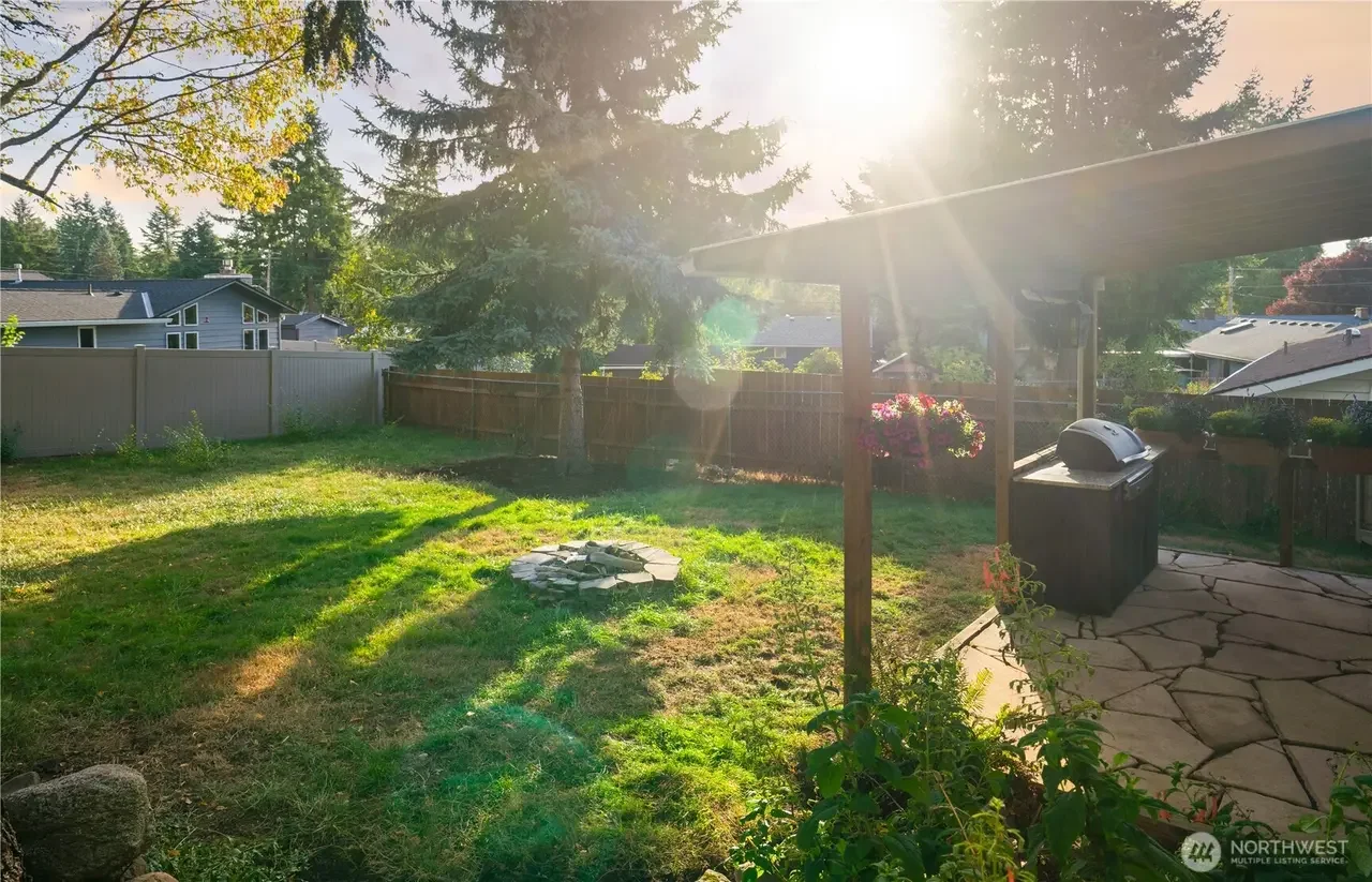 Backyard with green grass, multiple trees, a fenced perimeter, a covered patio area with a grill, flower pots, and the sun shining brightly with sun rays visible.
