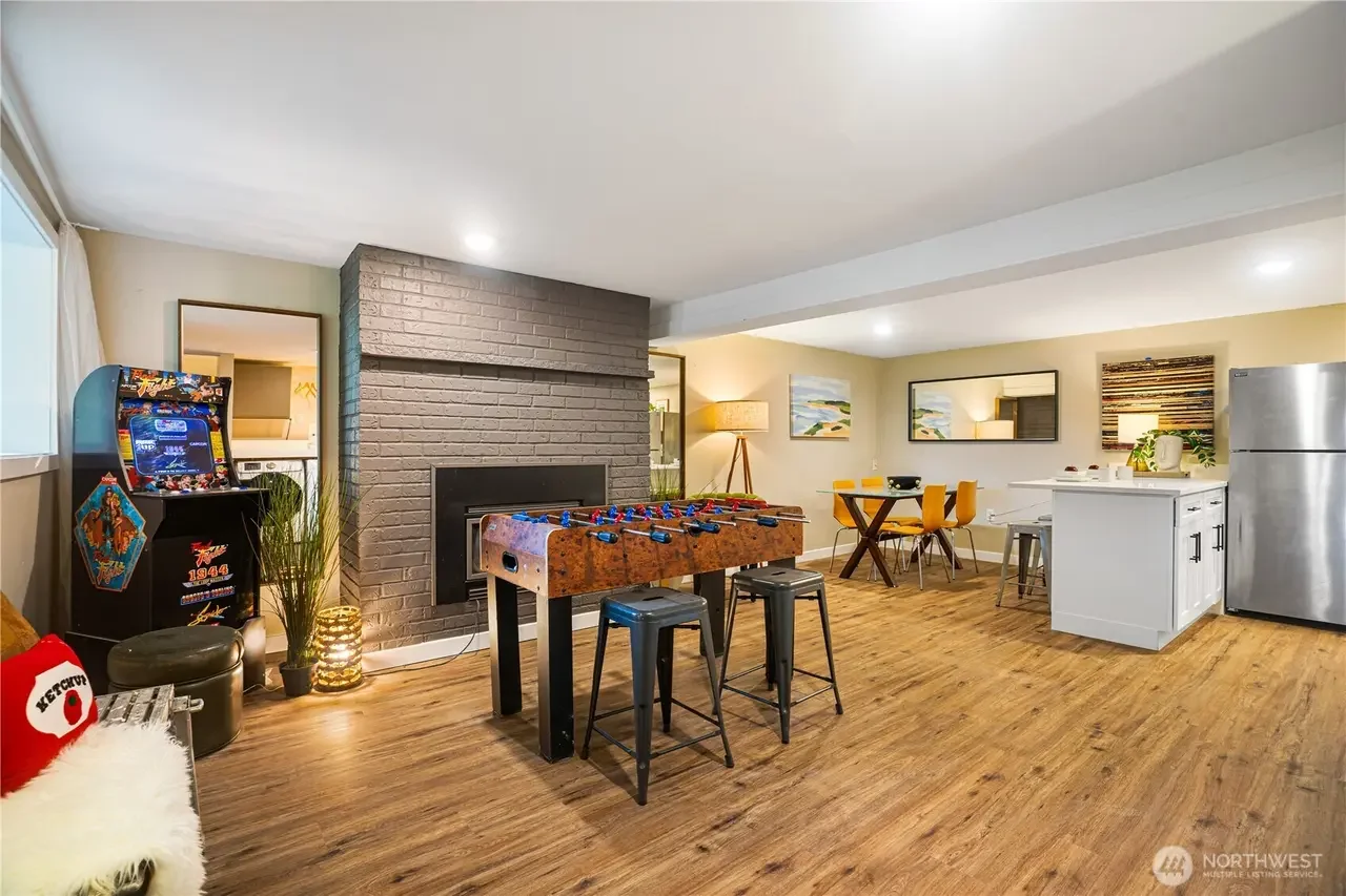 A spacious open-concept living room and kitchen with hardwood floors, a foosball table, a vintage arcade game, a fireplace, dining table with chairs, kitchen island, and stainless steel refrigerator. Decorative elements include paintings, a lamp, and