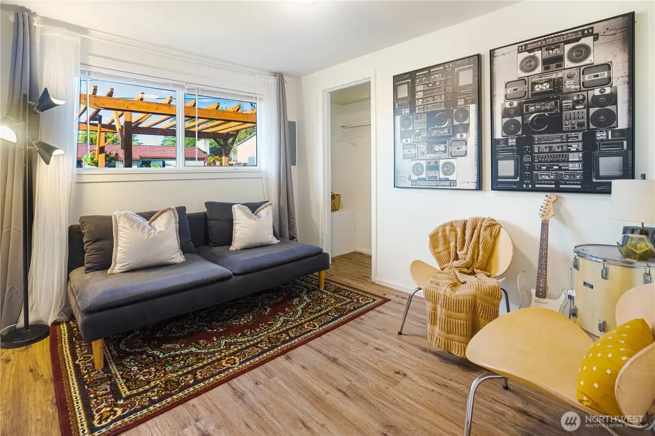 A living room with a gray sofa, white pillows, a black floor lamp, and a window with white curtains revealing a wooden pergola outside. There are two large black and white framed art pieces of boomboxes on the white wall. Two yellow chairs with pillo