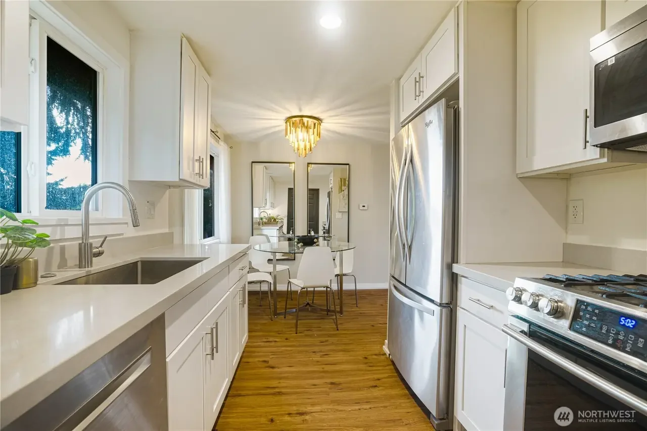 Modern kitchen with white cabinets, stainless steel appliances, hardwood floors, a round glass dining table with white chairs, and a decorative chandelier.