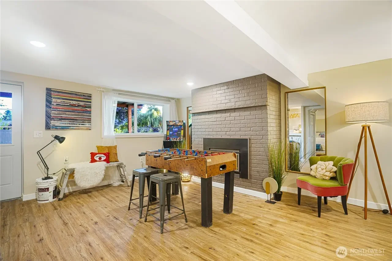 A cozy living room with a foosball table, colorful chair, tall mirror, floor lamp, and window with white curtains, overlooking a garden.