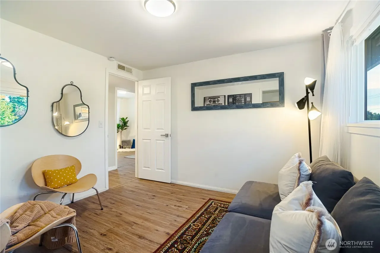Living room with a gray sofa, white pillows, a floor lamp, a window with curtains, two decorative mirrors on the wall, and a wooden floor.