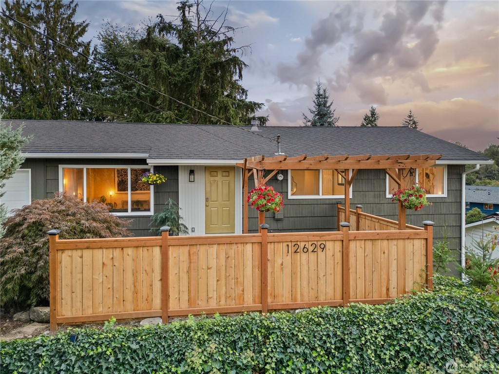 A single-story house with a green exterior, a gray shingled roof, and a small front porch with a wooden trellis. The yard includes a newly built wooden fence with hanging flower pots, plants, and shrubs in front and to the sides of the house.