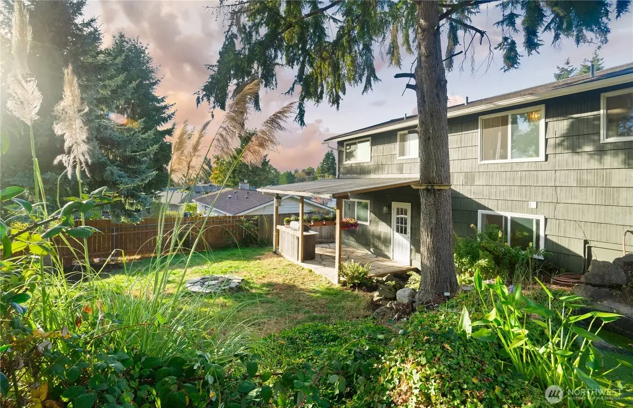 Backyard view of a two-story house with a covered patio, surrounded by trees and lush greenery, under a partly cloudy sky