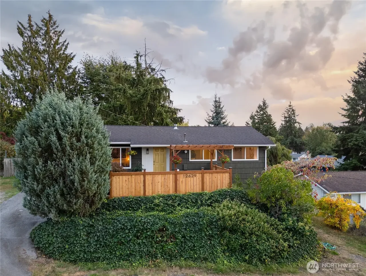A house with dark gray siding, a gray roof, and a wooden fence. The house features a small porch with hanging flower baskets. The yard has lush greenery, shrubs, and trees, with a cloudy sky overhead during sunset.