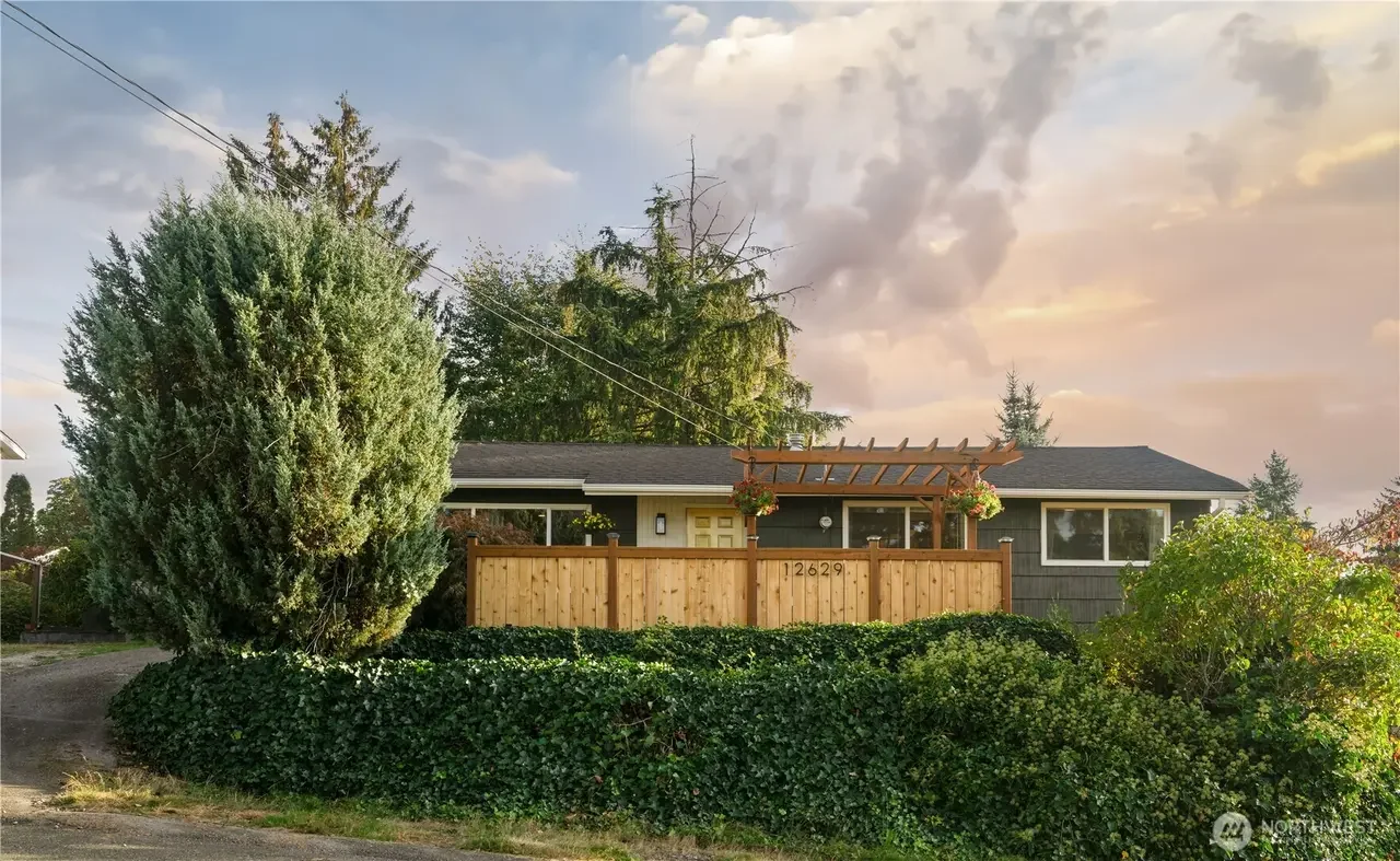 Single-story house with gray exterior and black roof, surrounded by trees and bushes, with a wooden fence and a small pergola on the front porch, at sunset with a cloudy sky.