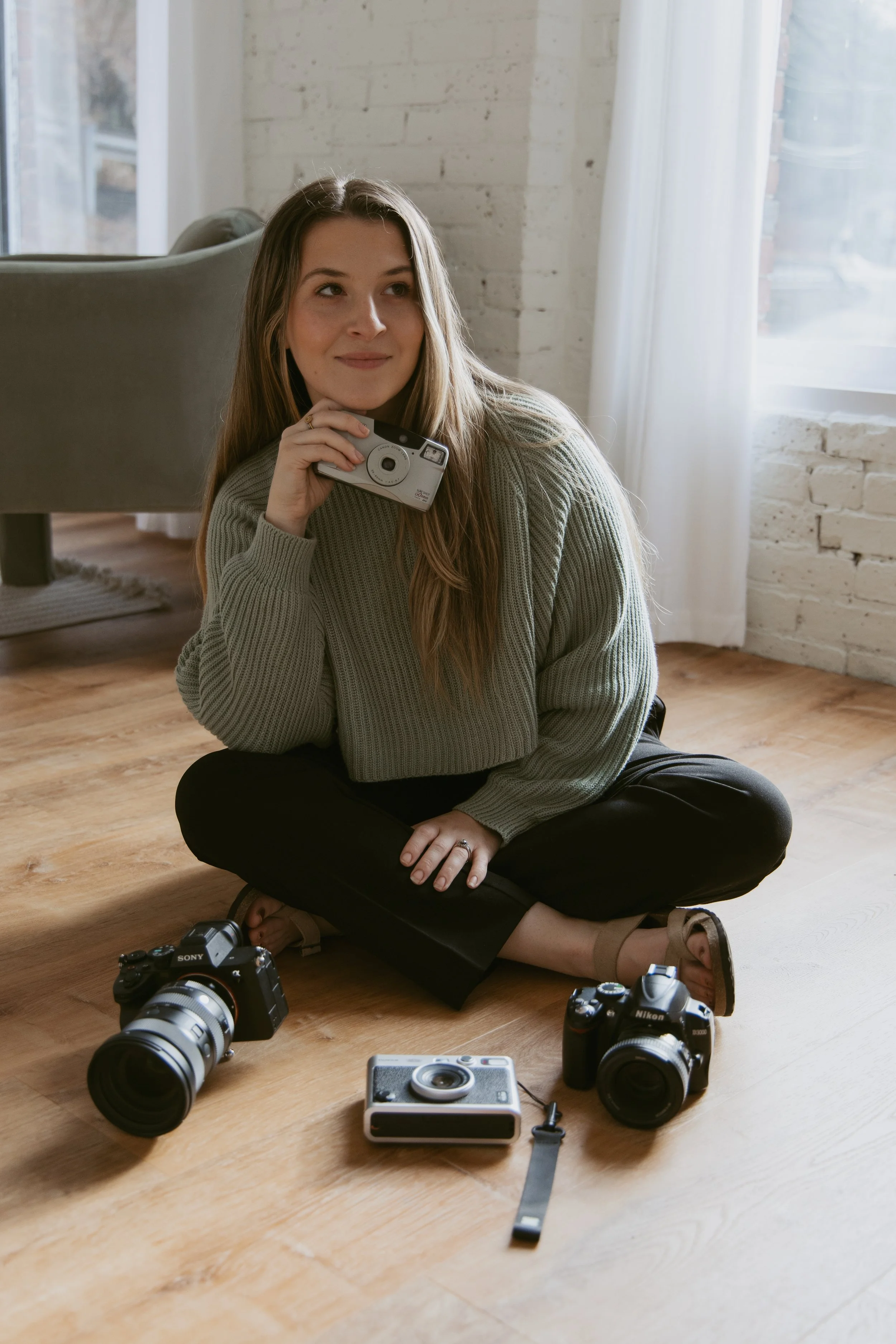 Young woman sitting on a wooden floor in a bright room with white brick walls, holding a camera near her face, surrounded by three other cameras and a small device with a strap.