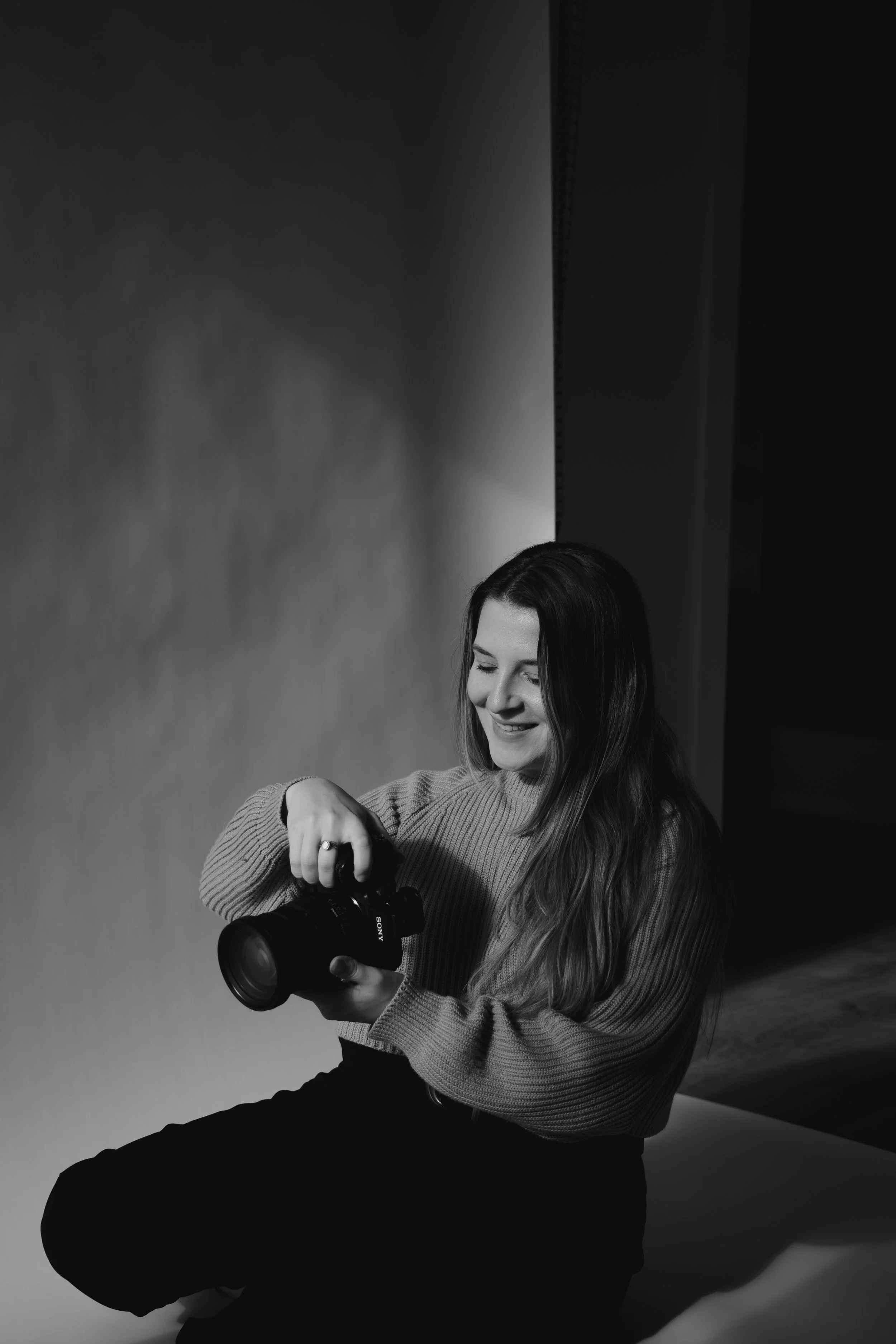 A woman with long hair sitting on the floor, holding a camera and smiling, in a light-colored sweater and dark pants, against a plain backdrop.