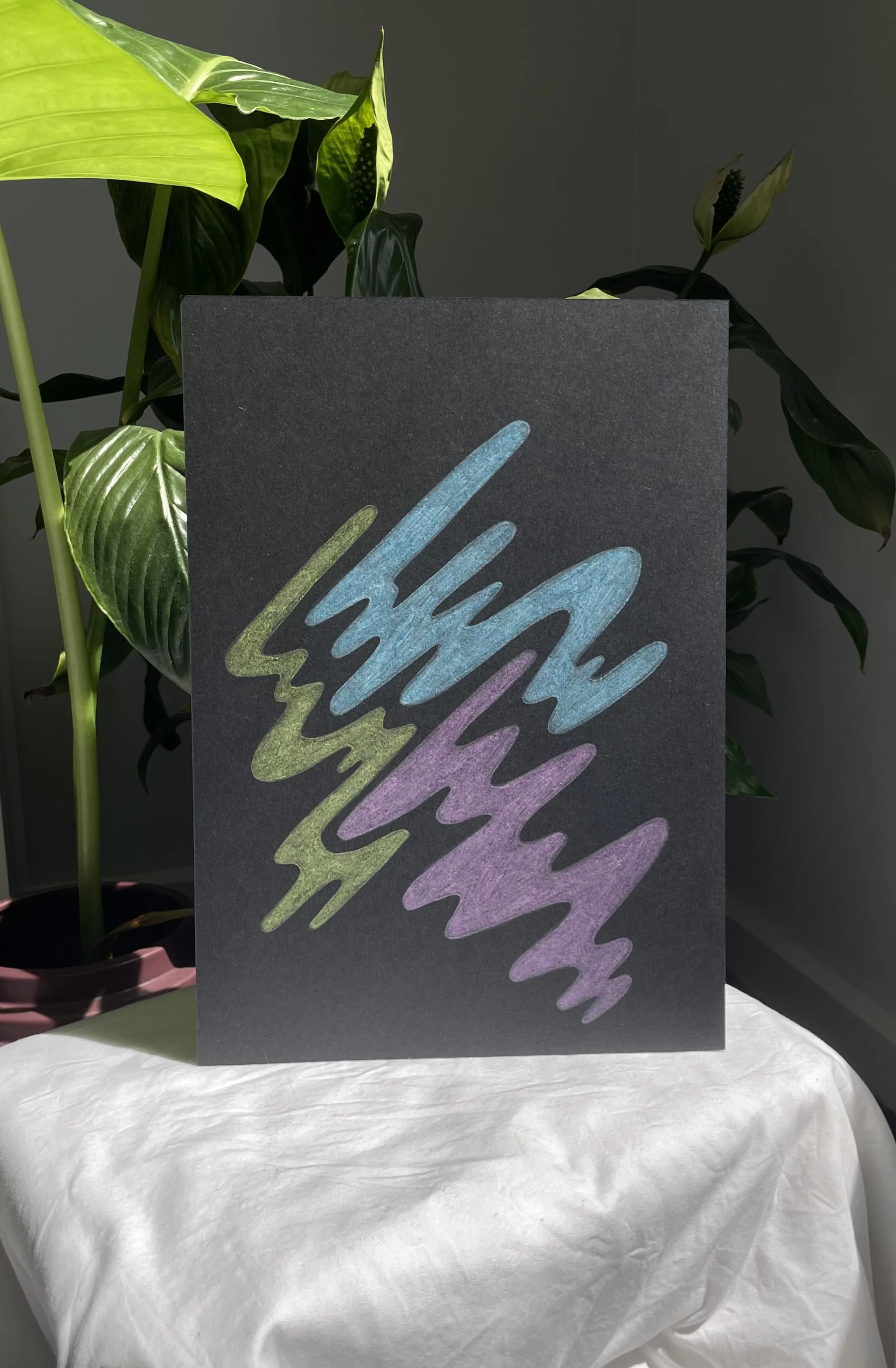 A dark-colored poster with colorful zigzag patterns in blue, purple, and green, placed on a table with a white cloth. Green leafy plants are in the background.