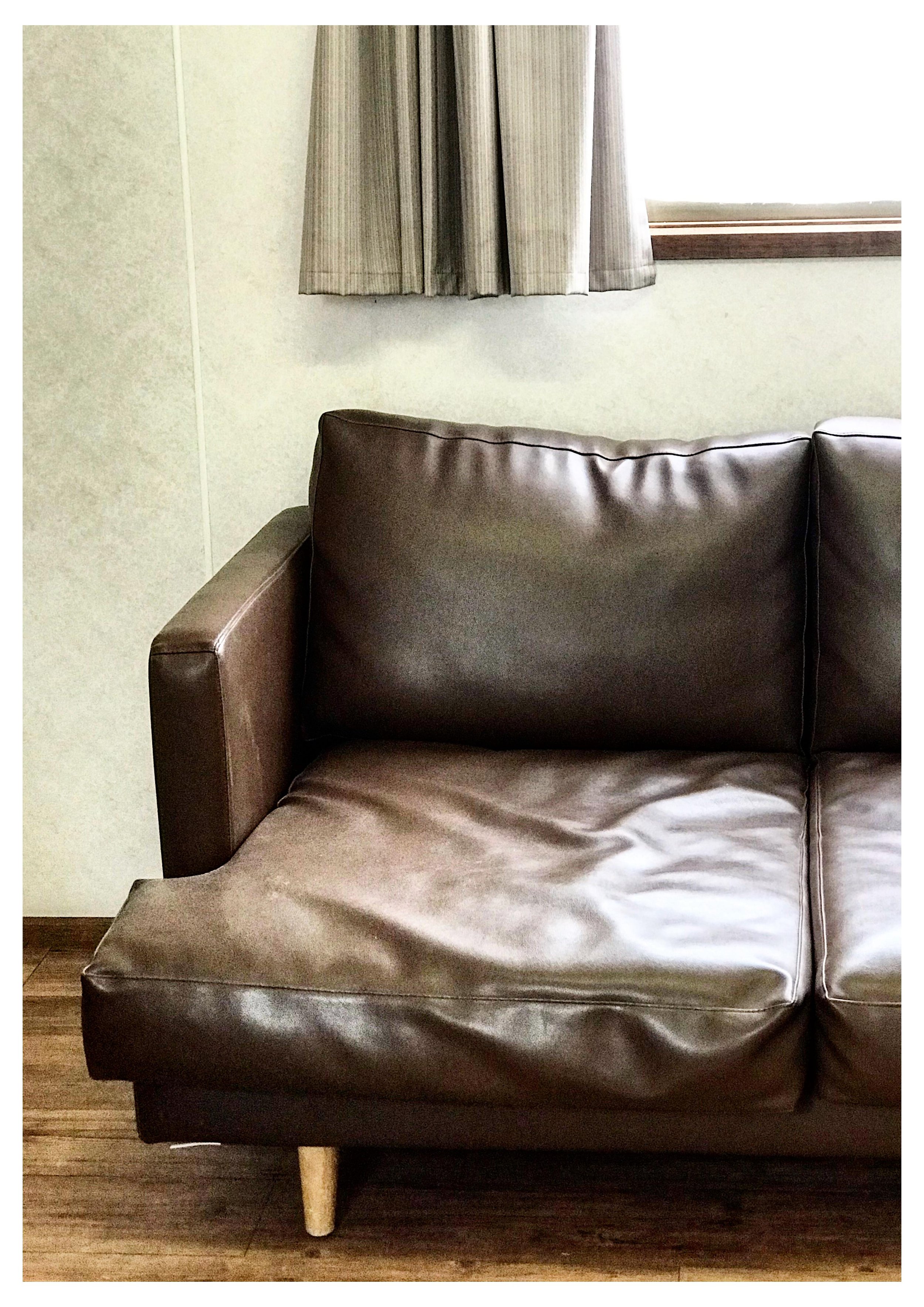 A brown leather couch with a cushion, positioned against a wall beneath a window with beige curtains. The floor is wooden.