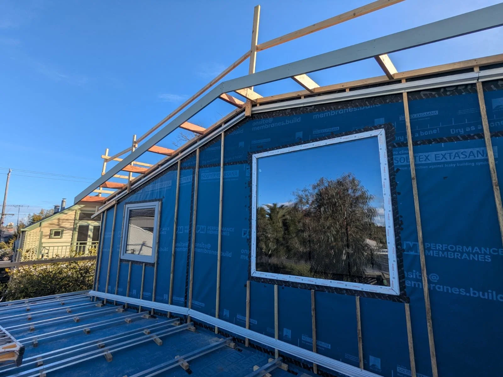 Construction of a building with blue weather-resistant sheathing and framed windows, with stairs in the foreground and a clear blue sky.