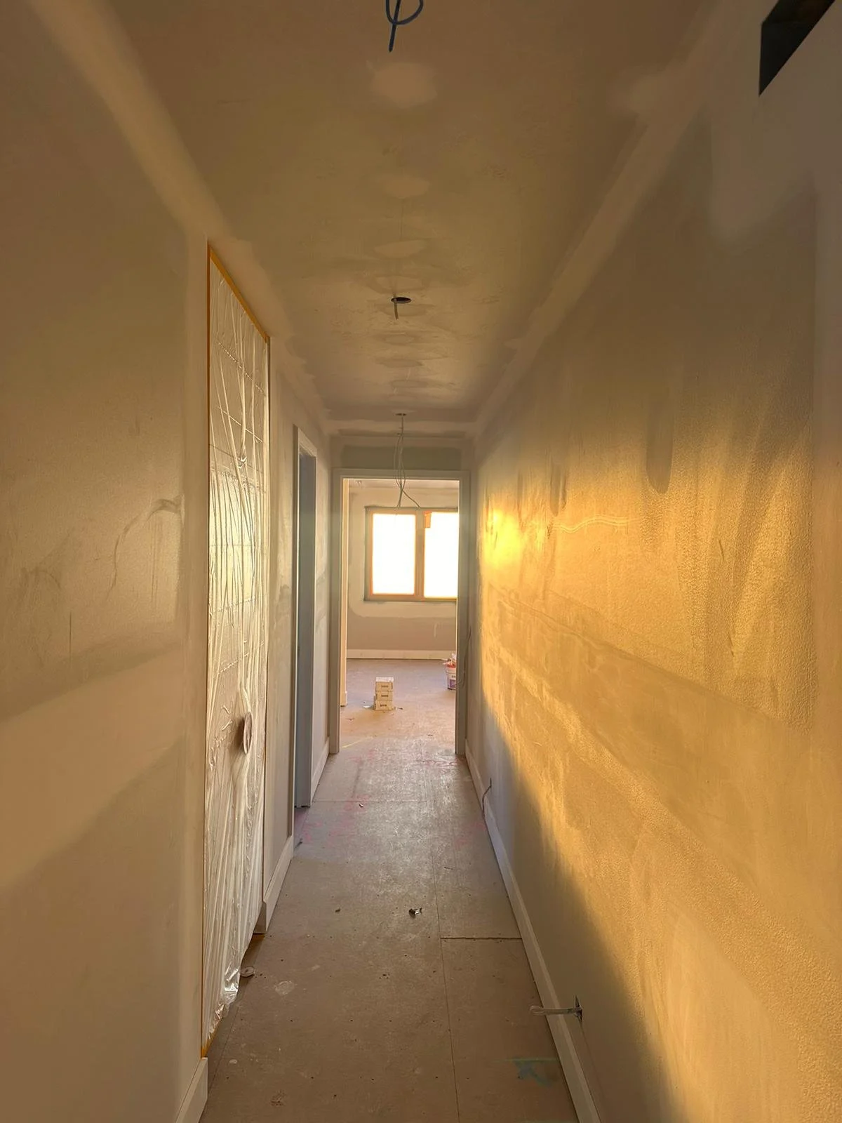 A hallway under construction with a window at the end, sunlight casting a warm glow on the walls, and construction materials on the floor.