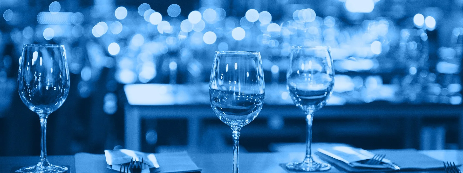 Three empty wine glasses on a table with folded napkins and utensils, with a blurred background of bokeh lights in blue tones.