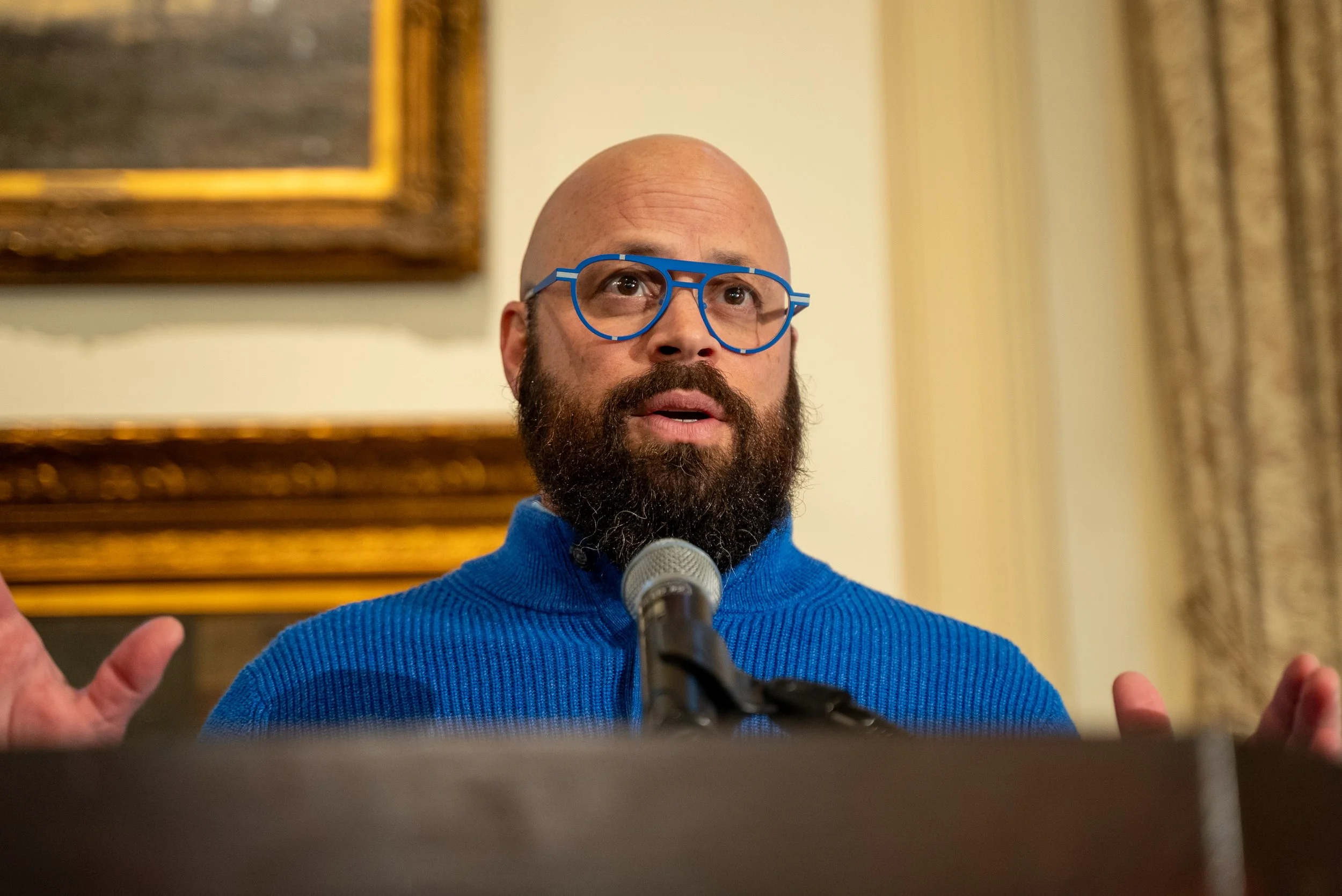 A man with a bald head and a beard, wearing blue glasses and a blue sweater, speaking into a microphone in a formal setting.