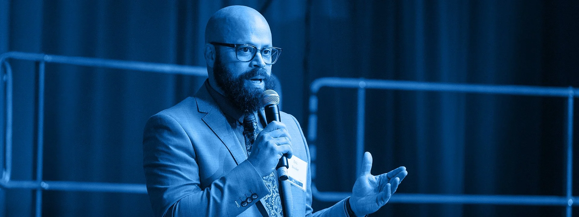 A man with glasses, a beard, and a bald head is wearing a suit and speaking into a microphone at a conference or event.