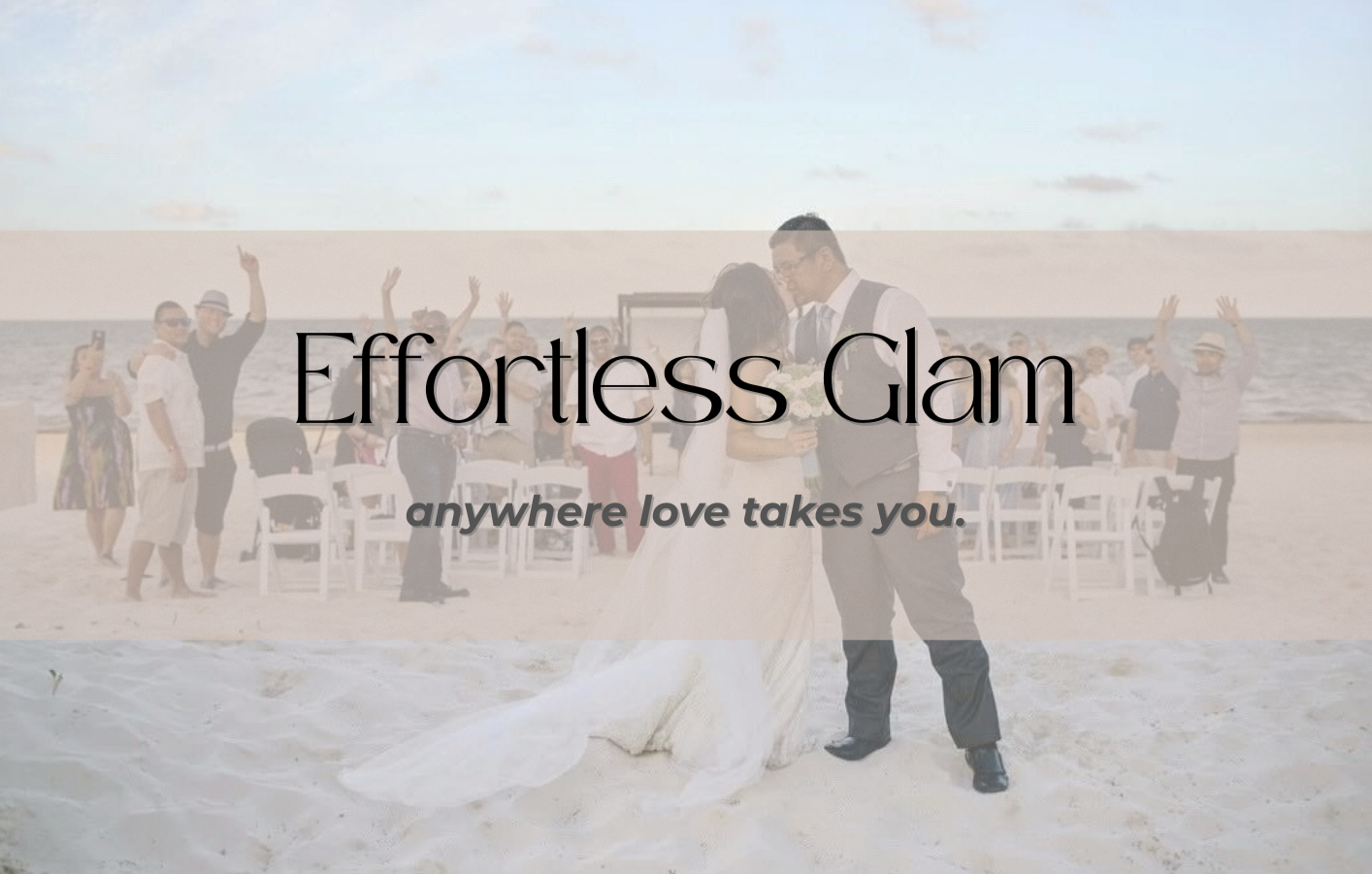 A couple in wedding attire embracing on a beach during their wedding ceremony with guests in the background, some taking photos and others raising their hands.