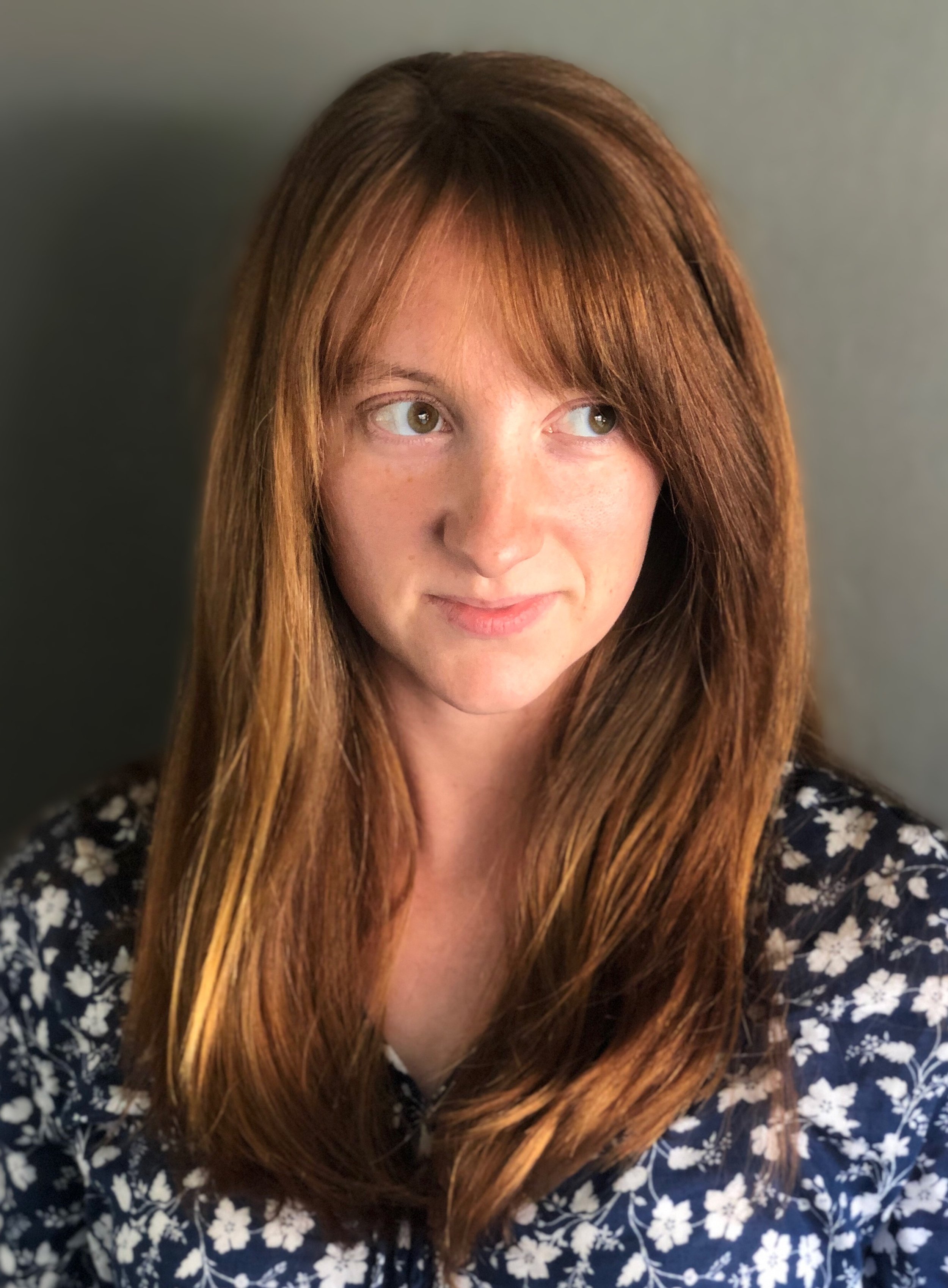 Woman with long brown hair, wearing a floral top, looking to the side.
