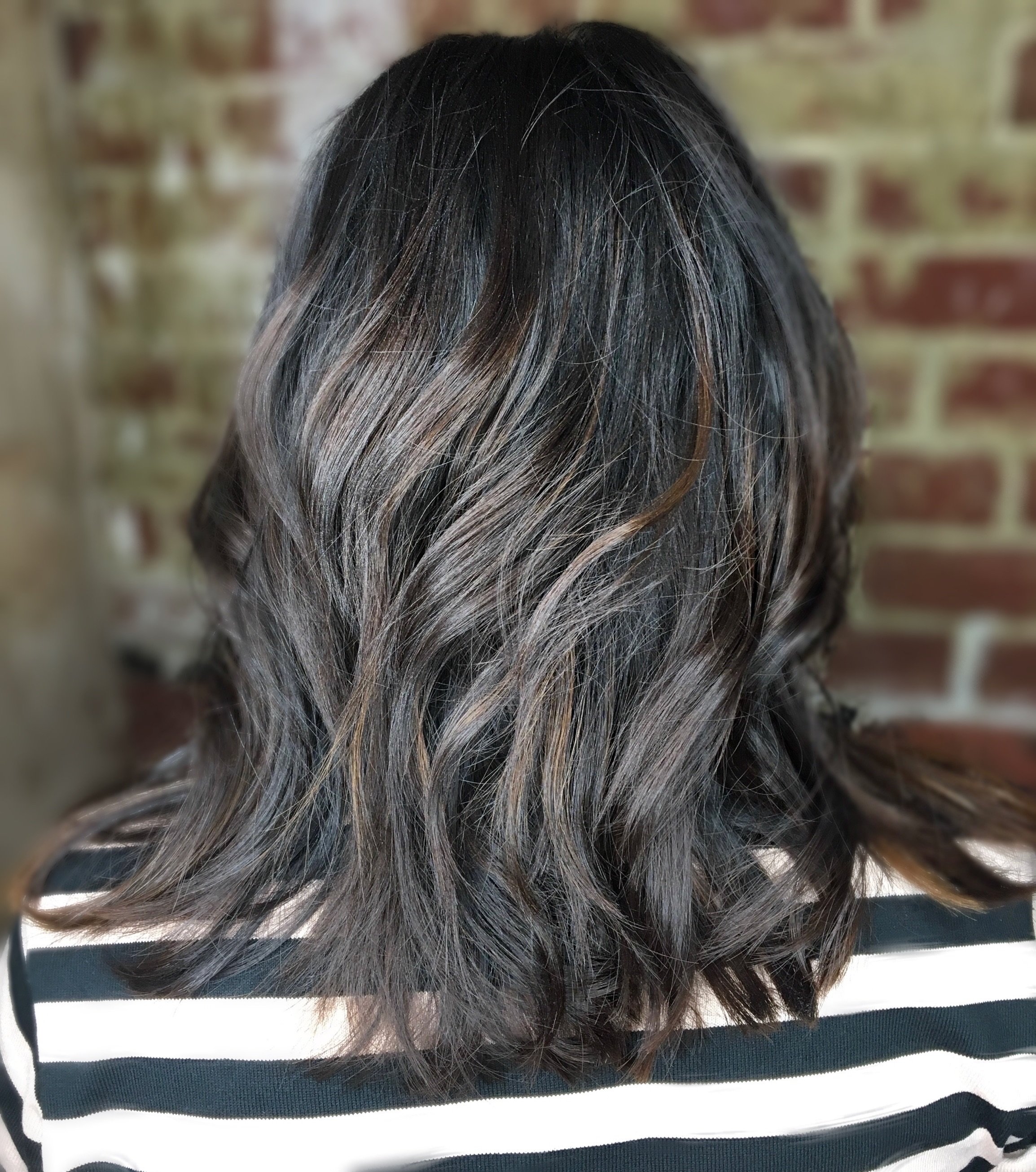 Back view of a person with dark, wavy shoulder-length hair against a brick wall background.