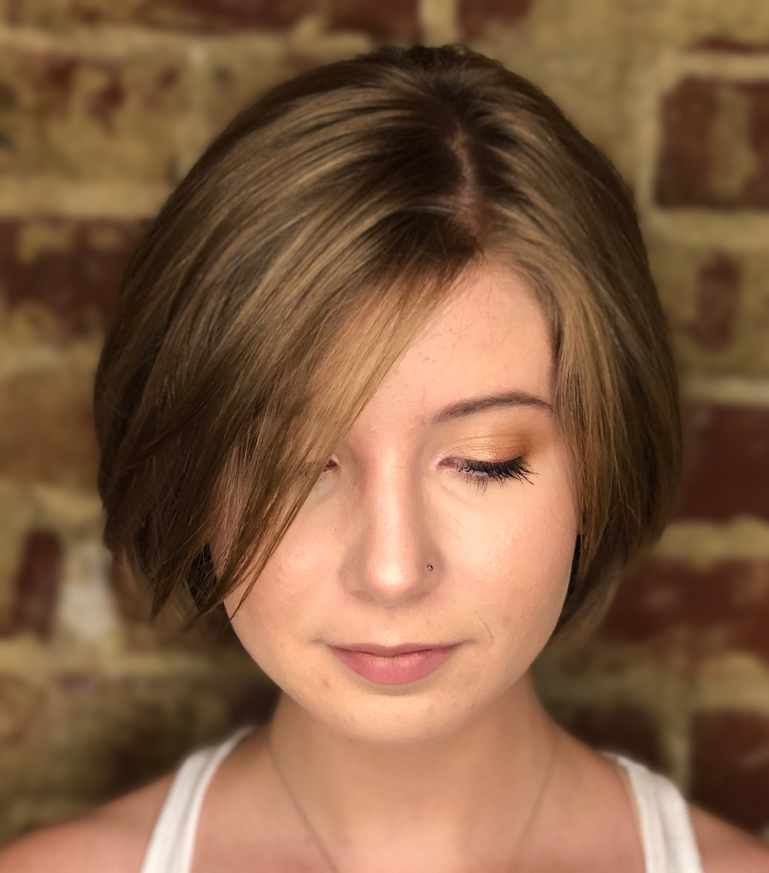 Woman with short brown hair and a nose piercing, wearing a white top, against a brick wall background.