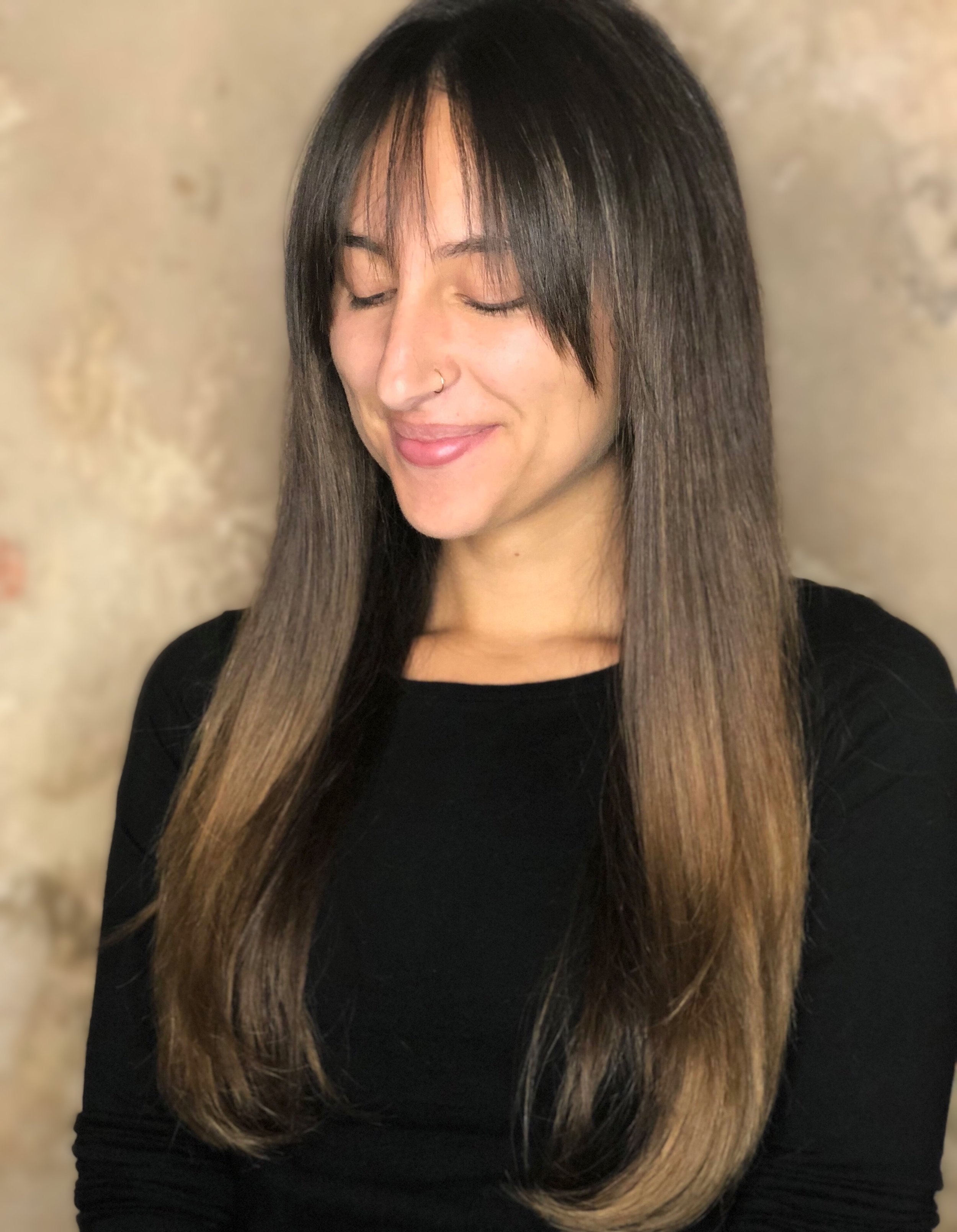 Person with long straight brown hair and bangs, wearing a black shirt, smiling with closed eyes against a neutral background.