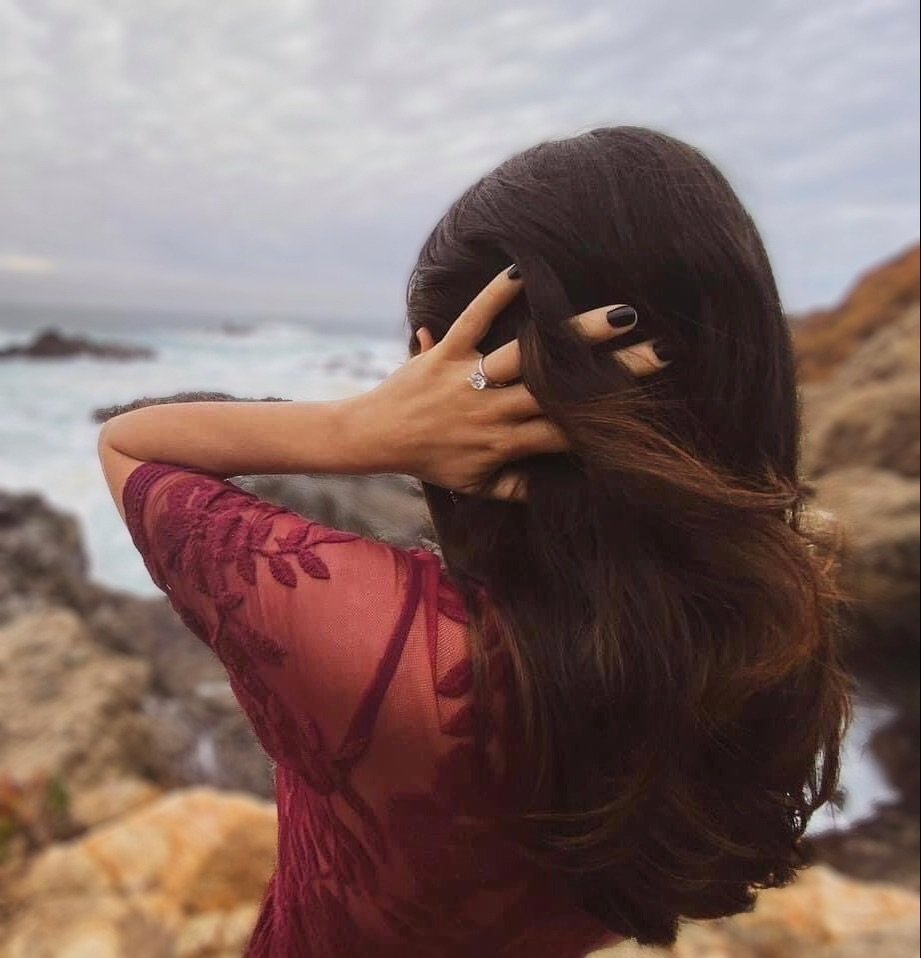 Woman in maroon dress with lace detail stands by rocky coastal scene, hand in hair revealing a ring.