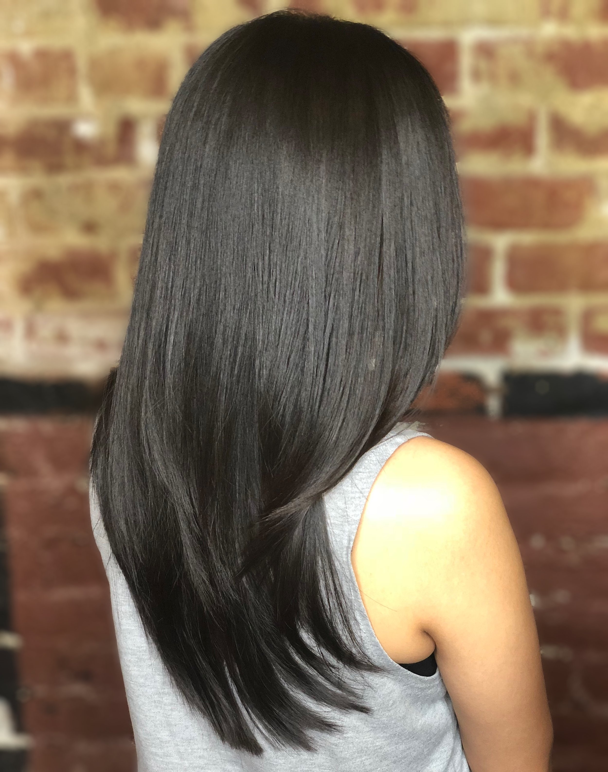 Back of a woman with long, straight, black hair wearing a gray tank top, brick wall background.