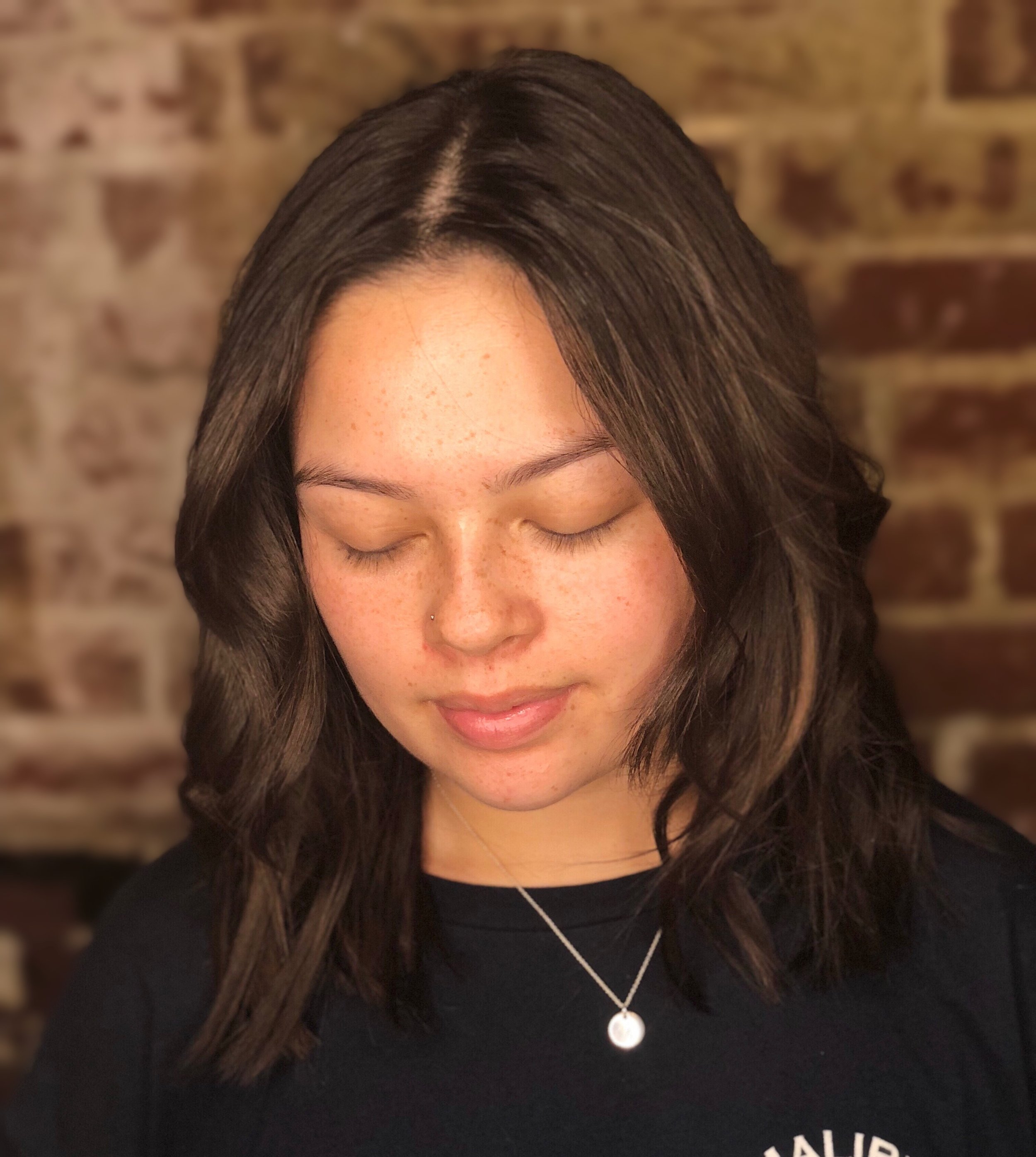 Young woman with closed eyes, wearing a necklace, against a brick wall background.
