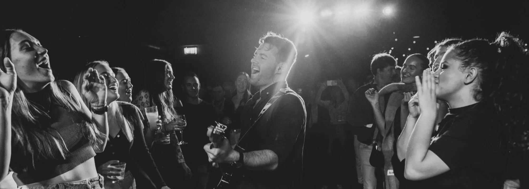 A group of people at a party or concert, enjoying music and dancing, with a man playing guitar at the center and everyone smiling and singing along.