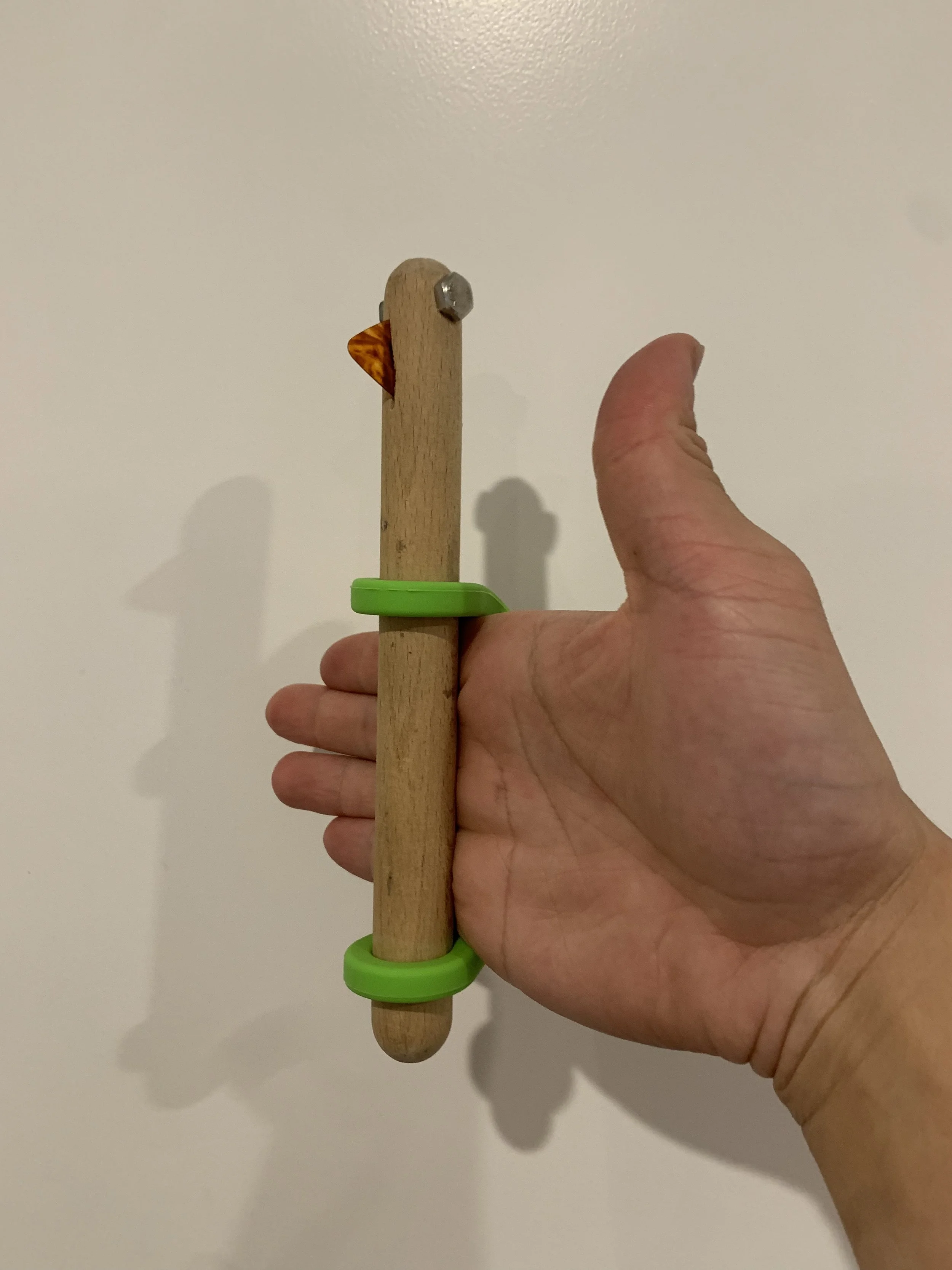 Hand holding a wooden chicken toy with an orange beak and a metal bolt eye, secured with two green rubber bands.