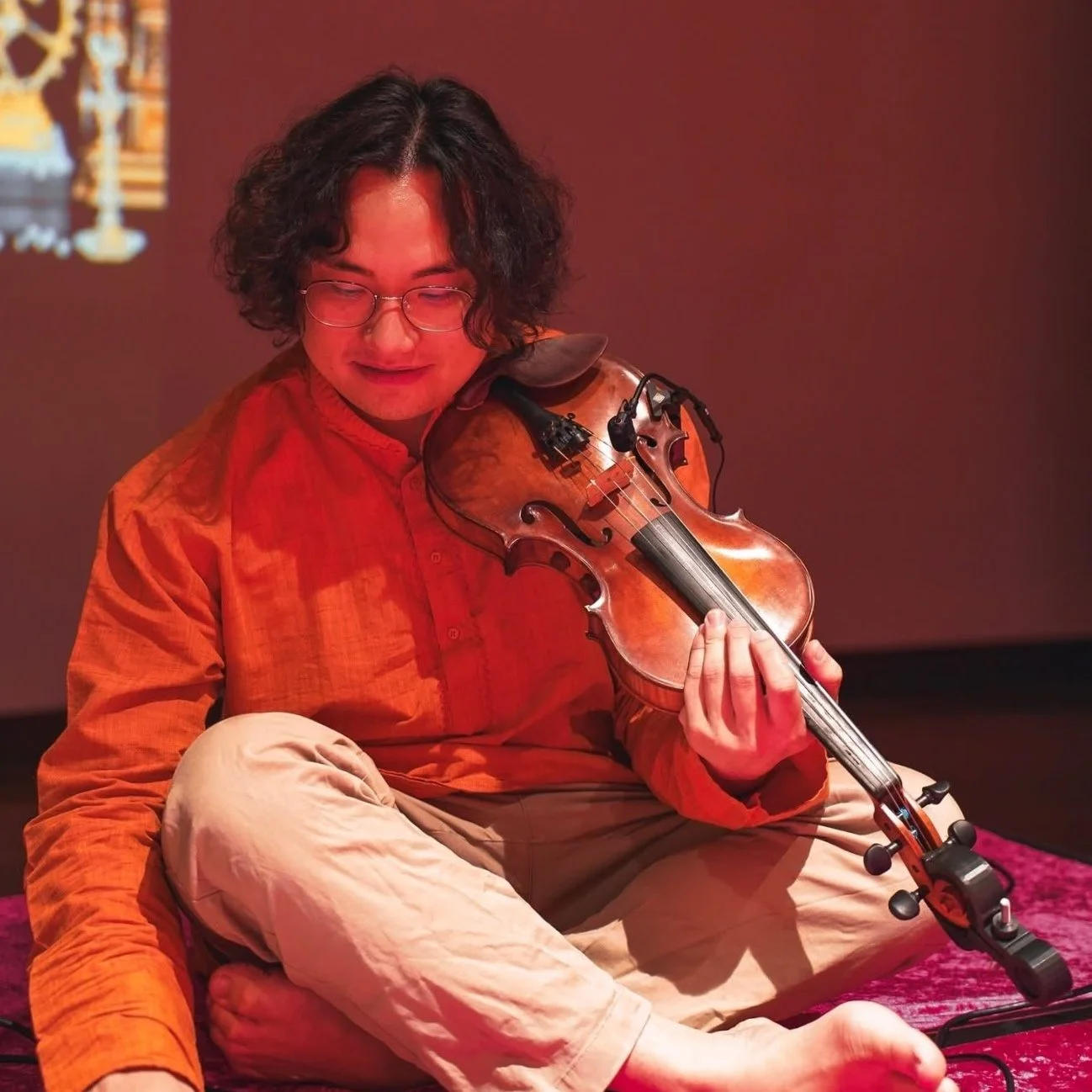 Person with curly dark hair and glasses sitting cross-legged, playing a violin, wearing an orange shirt and beige pants.