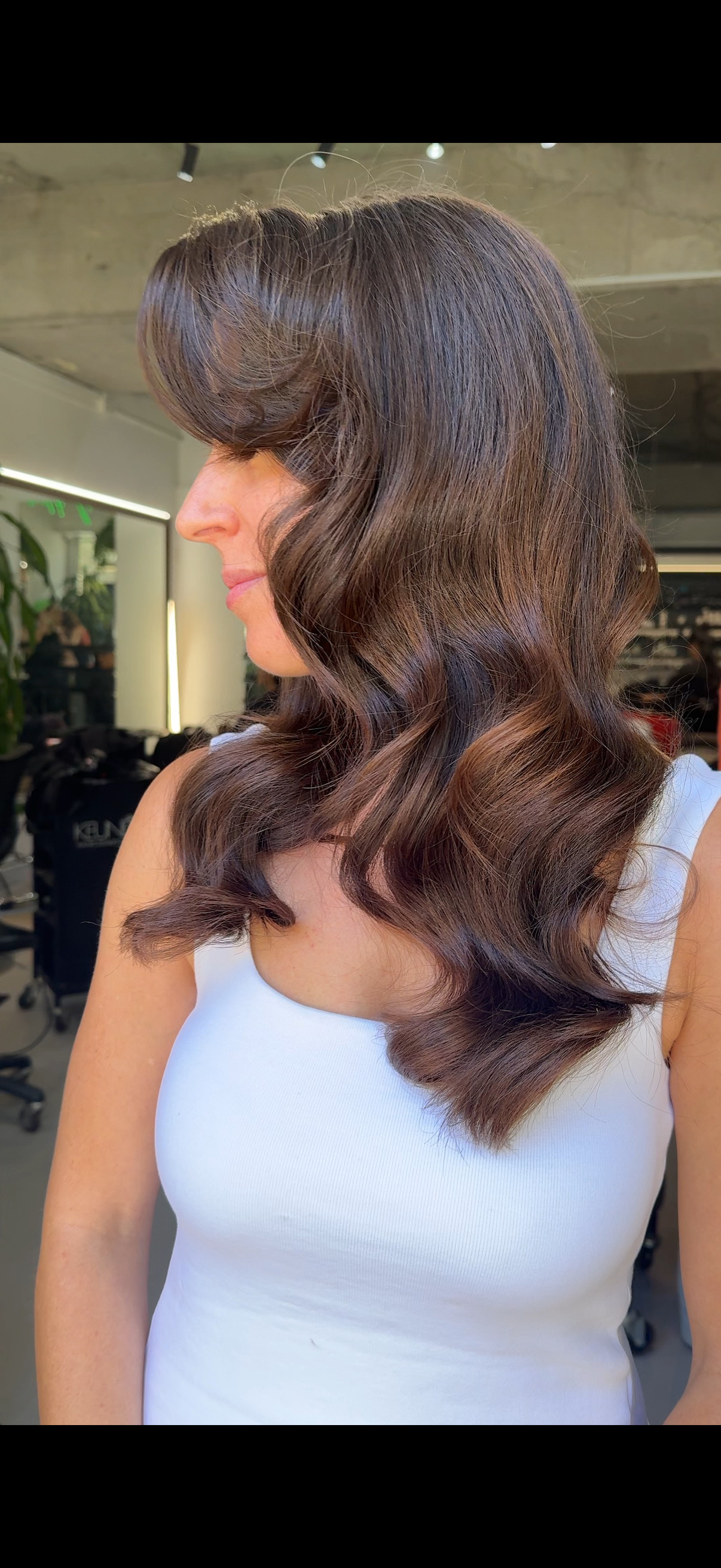 A woman with long, wavy brown hair and makeup with eyeshadow and lipstick, posing against a neutral background.