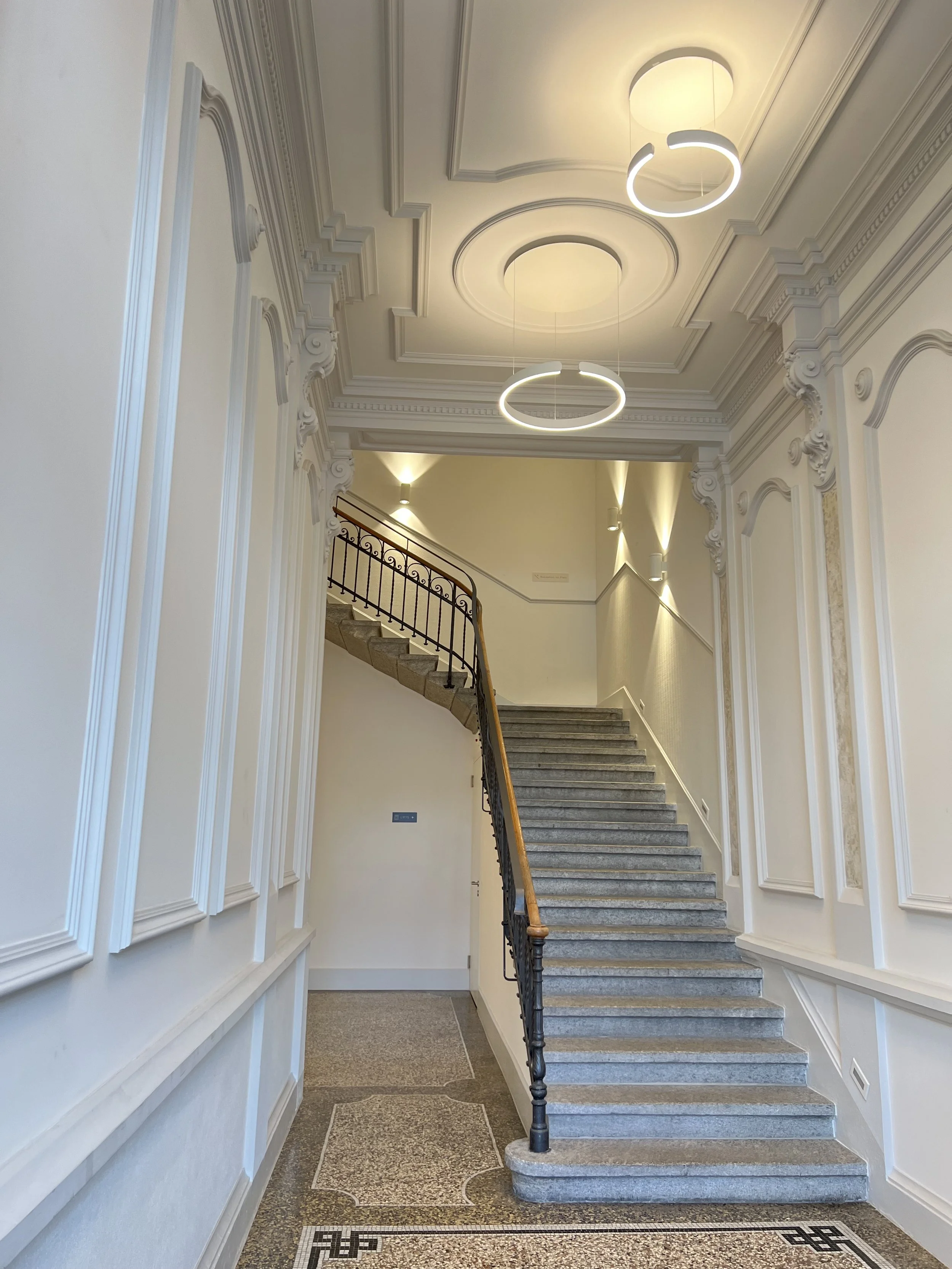 Interior staircase with ornate molding, circular ceiling lights, and wrought iron railing.