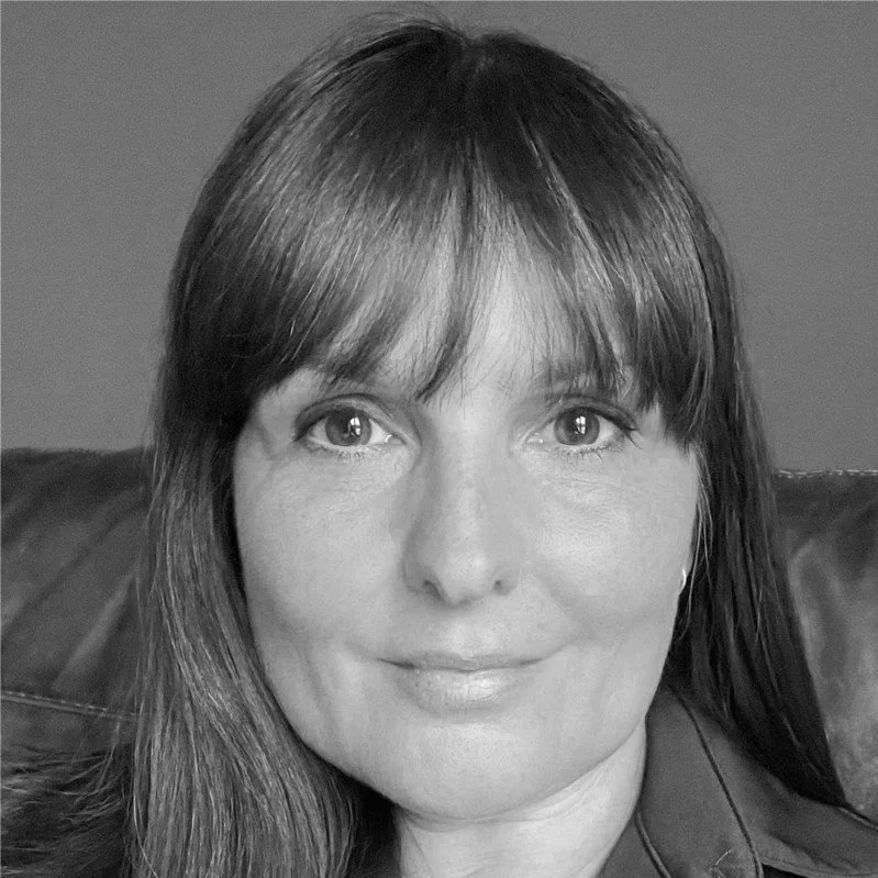 Close-up black and white photo of a woman with long dark hair, wearing a collared shirt, sitting on a leather sofa with a plain background.