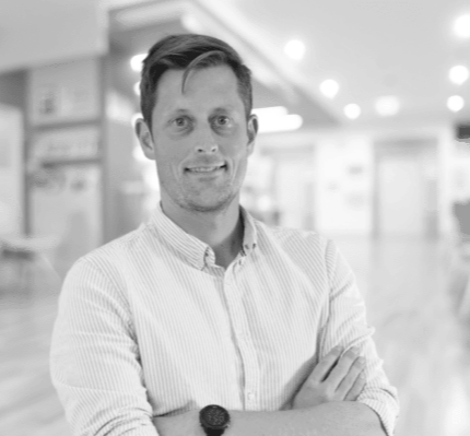 A man with dark hair, wearing a striped button-up shirt, standing indoors with his arms crossed, smiling at the camera in a modern, well-lit space.