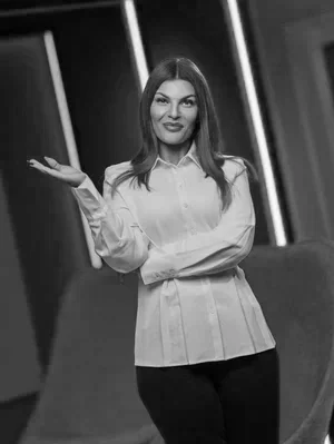 A woman standing indoors with a confident smile, wearing a white blouse and black pants, with her arms crossed and one hand gesturing outward.