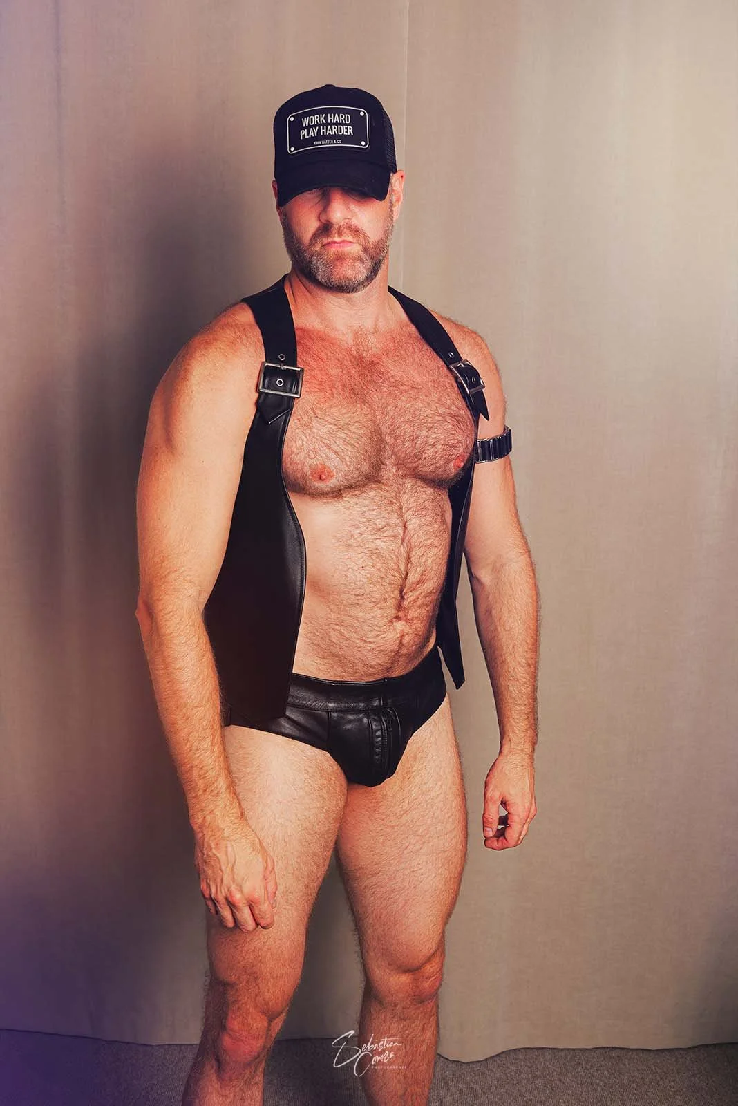 Man posing topless, wearing a black cap with 'Work Hard Play Harder' message, leather vest and shorts, with a plain background.