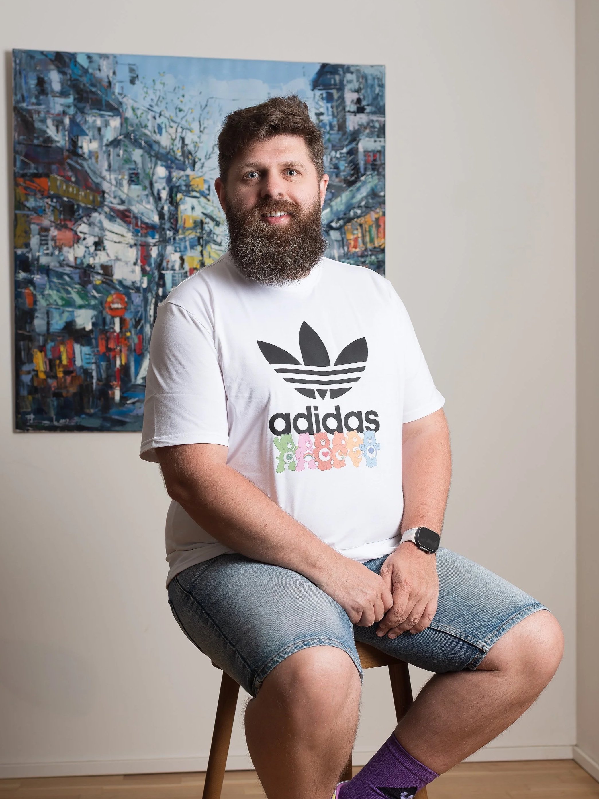 A man with a beard and wearing a white Adidas t-shirt and denim shorts is sitting on a wooden stool. He has a smartwatch on his left wrist and is wearing purple socks. Behind him, there is a colorful painting of a city scene on a beige wall.