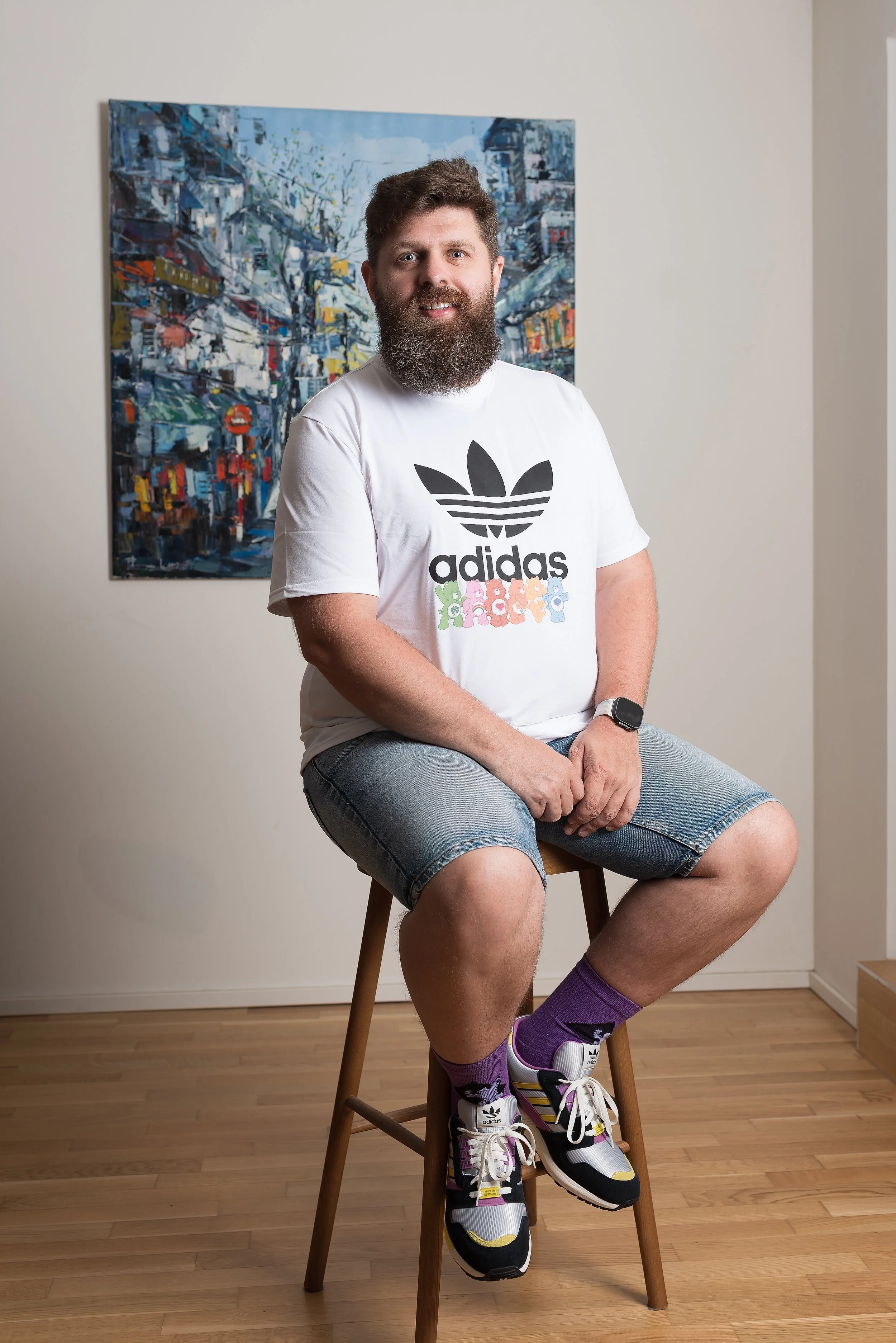A man with a beard sitting on a wooden stool in front of a colorful cityscape painting. He is wearing a white T-shirt with a black Adidas logo and rainbow-colored lettering, denim shorts, purple socks, and Adidas sneakers.