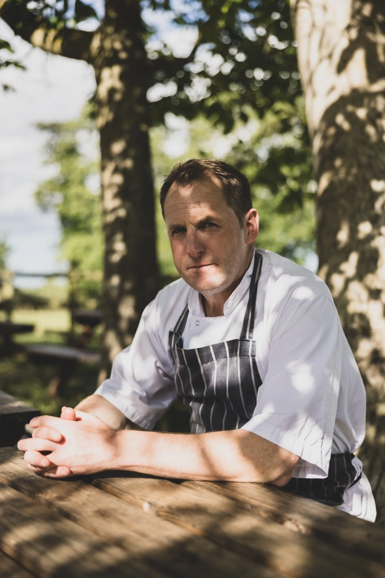 A male chef in a striped apron and white chef's coat sitting outdoors at a wooden table, with trees and sunlight in the background.