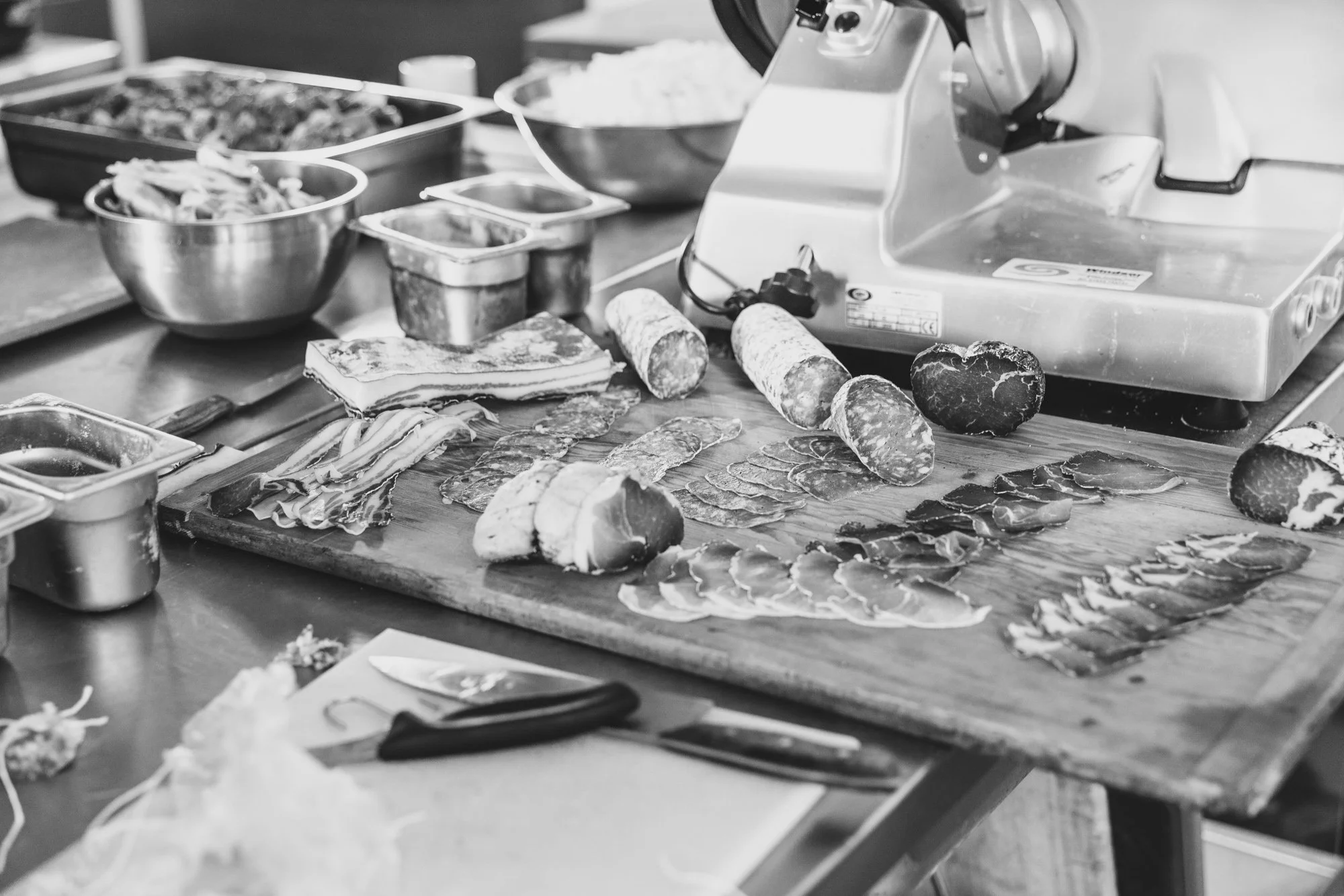 A kitchen countertop with sliced meats, sausages, and various ingredients, along with a meat slicer and cooking dishes in the background.